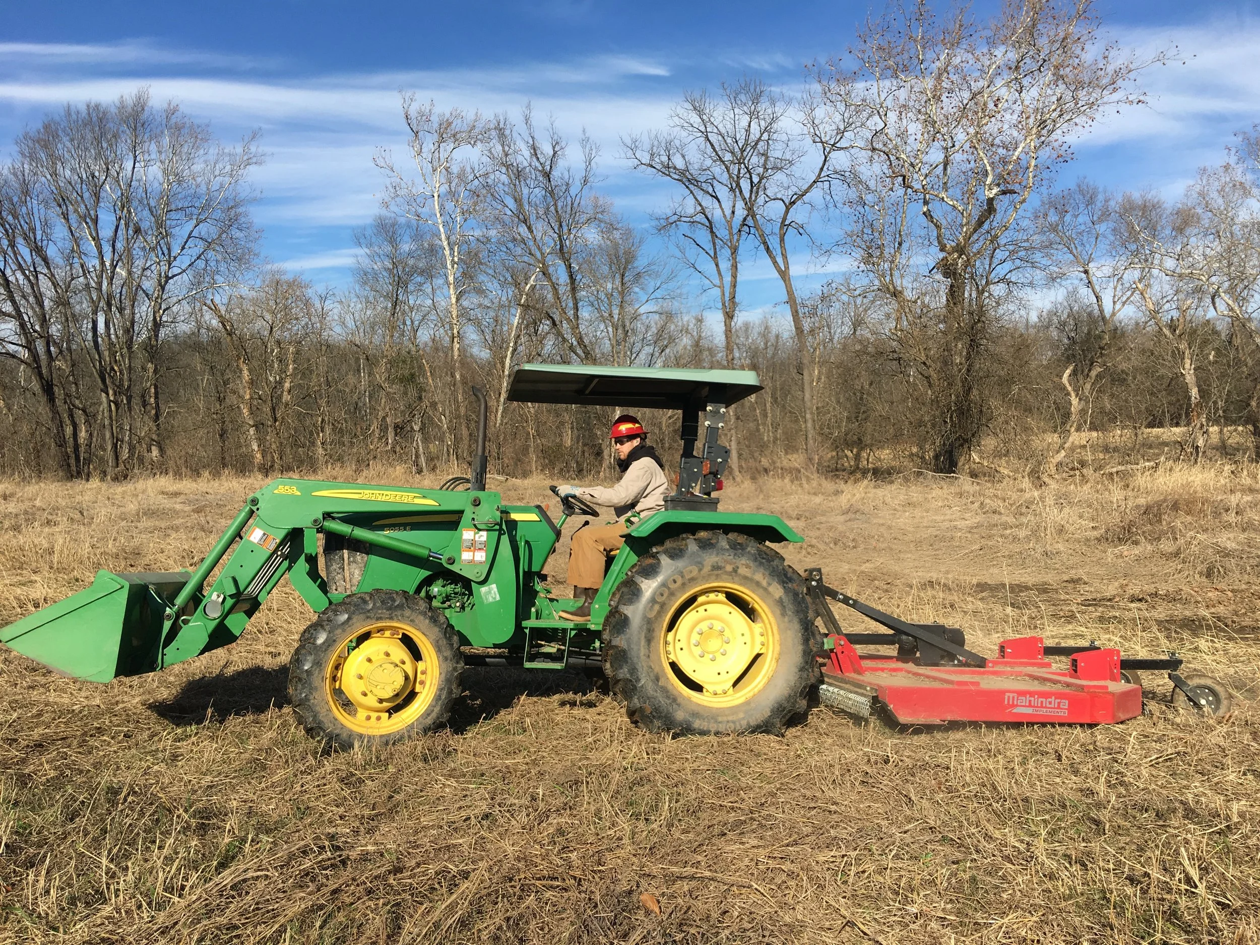A person wearing a red helmet operating a green tractor with a red mower attachment in a field with bare trees and blue sky.