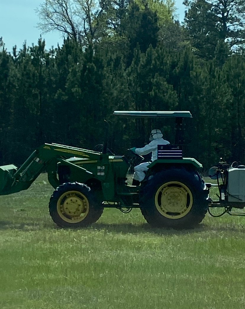A person dressed in white protective suit operating a green tractor with a sprayer in a grassy field with trees in the background.
