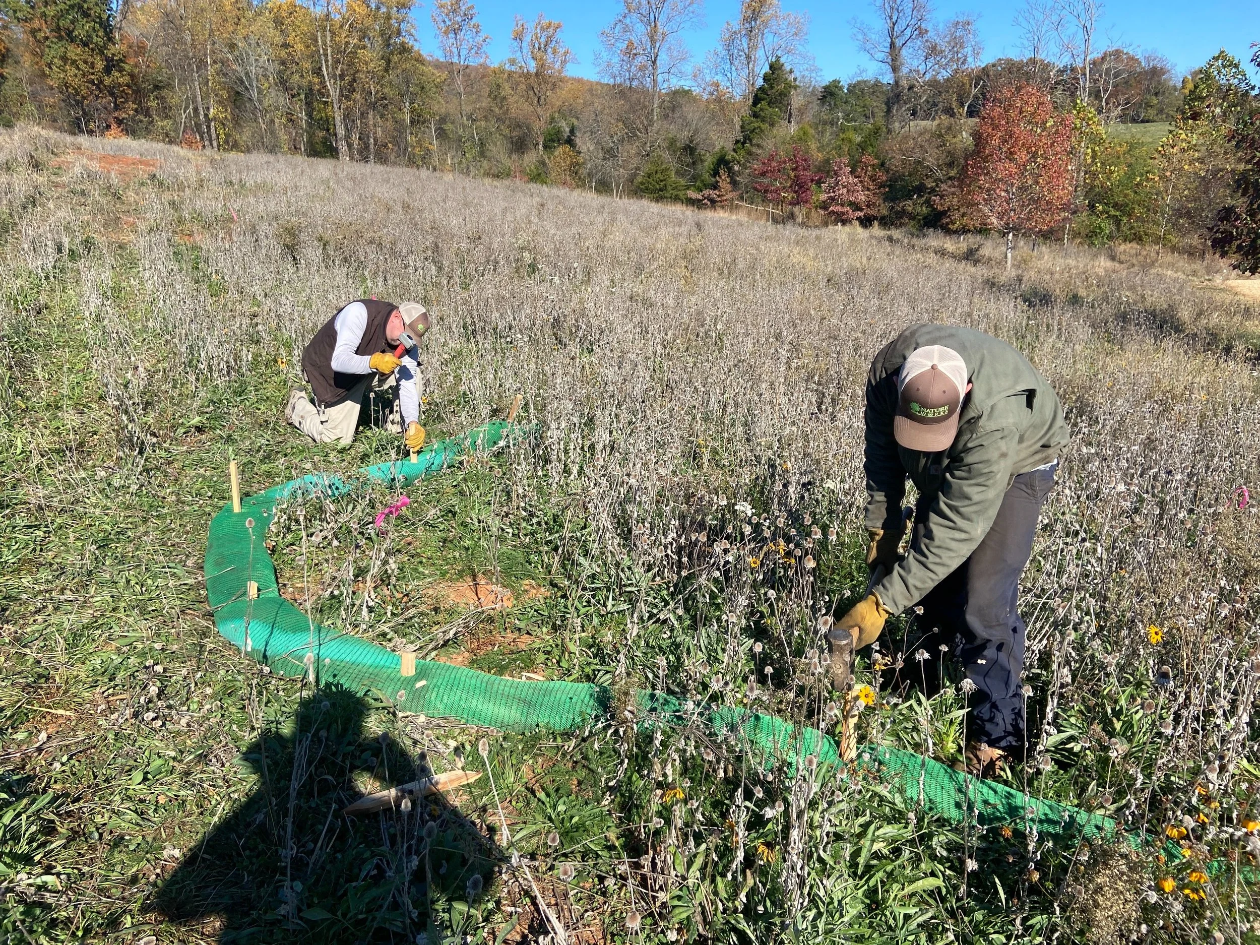 Two people working in a field of wildflowers with trees in the background, using tools near a green flexible barrier.