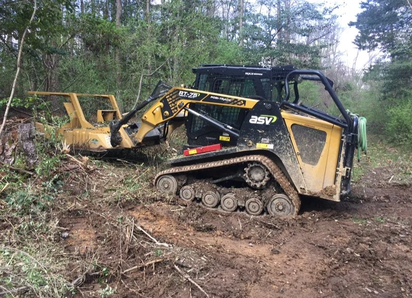 A small black and yellow tracked skid-steer loader with a hydraulic rake attachment clearing trees and debris in a wooded area.