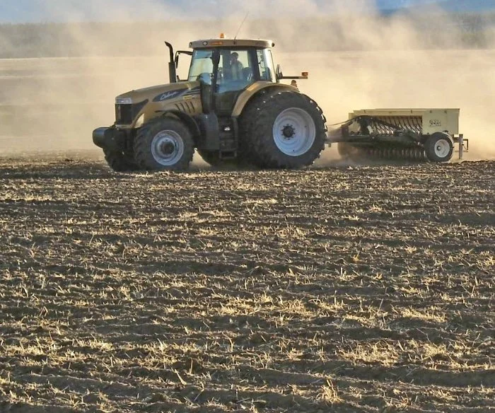 A tractor pulling a seed spreader across a dirt field, creating a trail of dust.