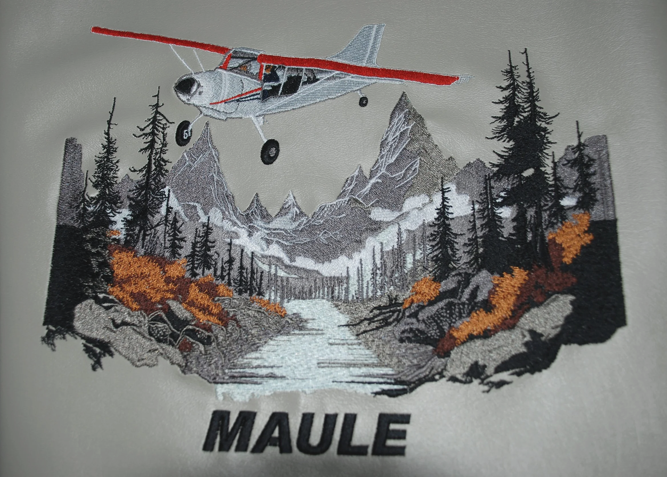 mountain seance with a maule airplane flying over it embroidered on a tan piece of leatherette.