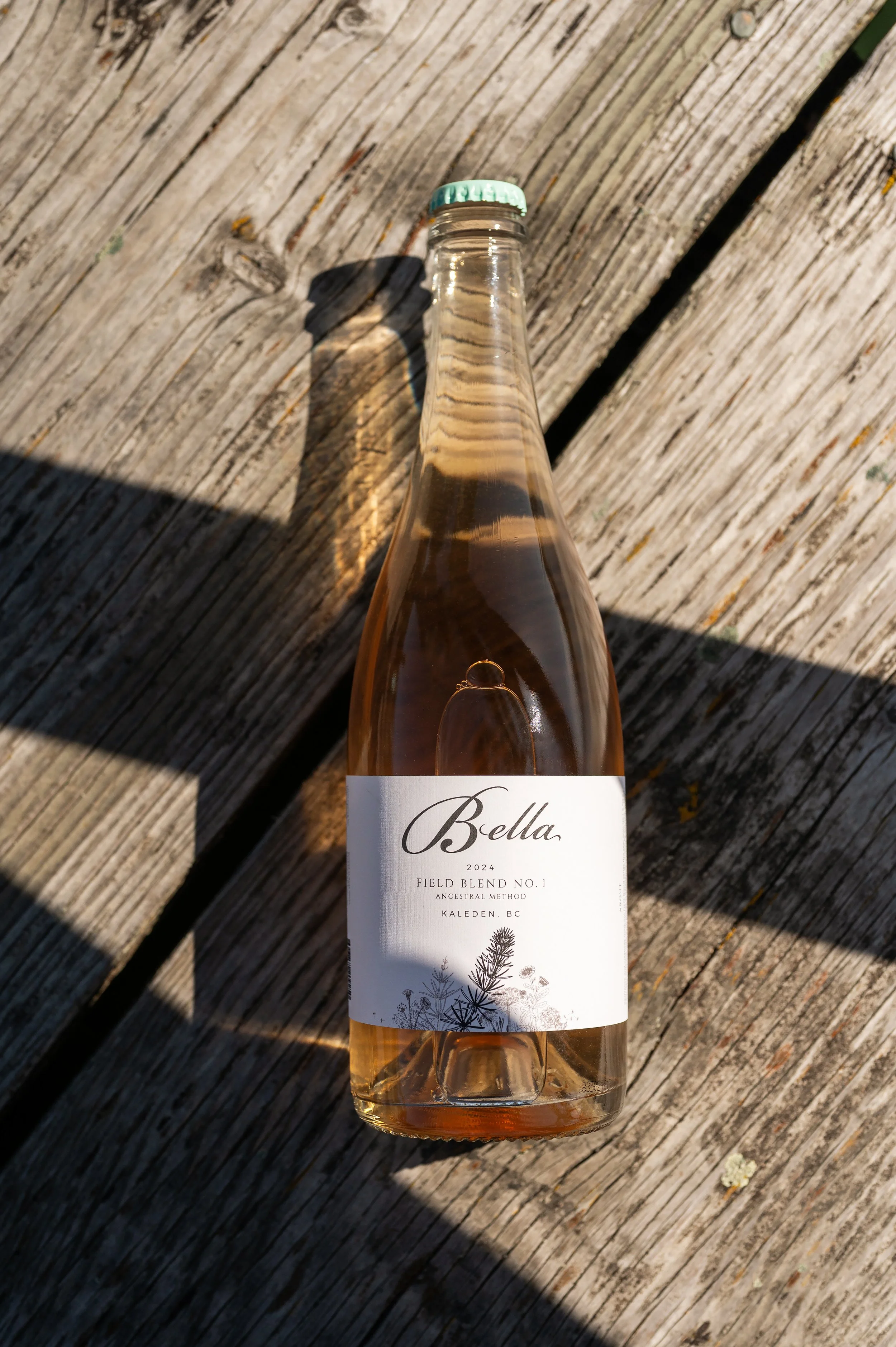 A glass bottle with a mint green cap, labeled 'Bella' with a botanical illustration and text indicating it is a 2024 field blend wine from Kaleden, BC, resting on a weathered wooden surface outdoors.