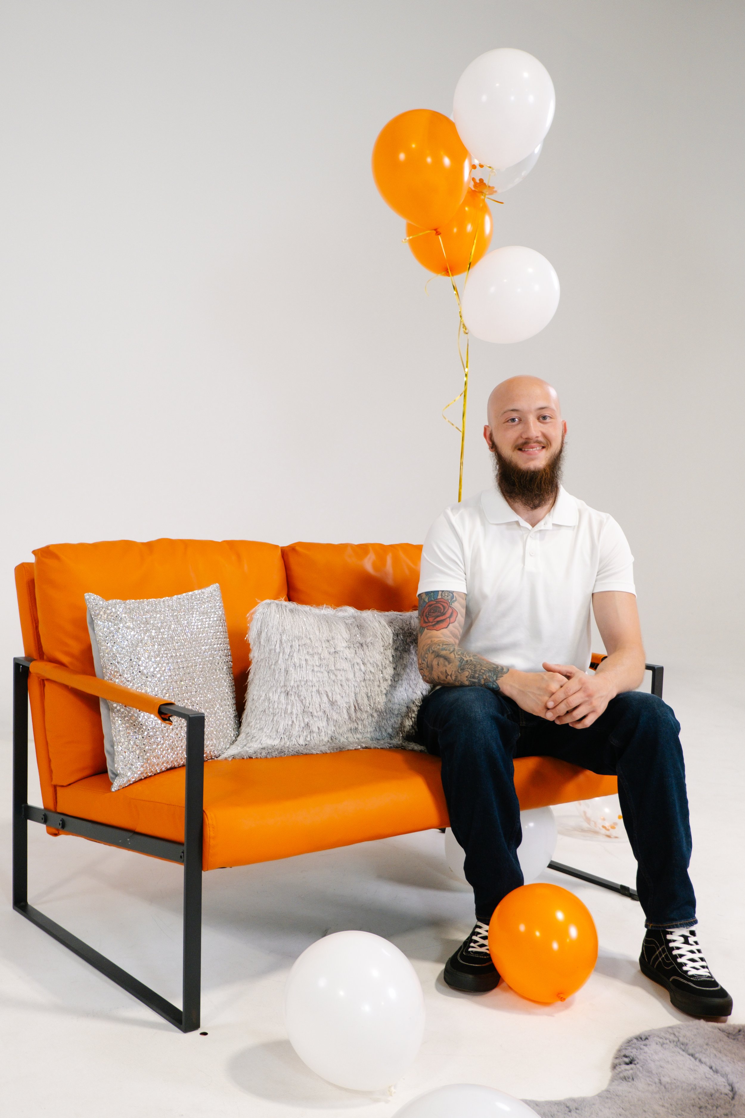 A smiling man with tattoos on his arm, seated on an orange sofa, surrounded by white and orange balloons, in a minimal white room.