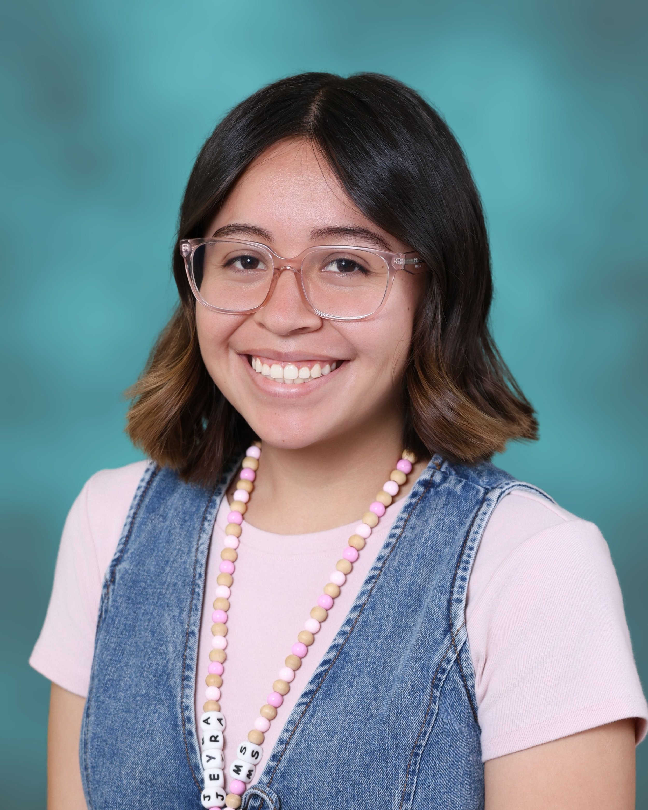 Close-up of a young woman with dark hair, wearing clear eyeglasses, a light pink t-shirt, a denim vest, and a beaded necklace with pink, white, and beige beads, smiling against a teal background.