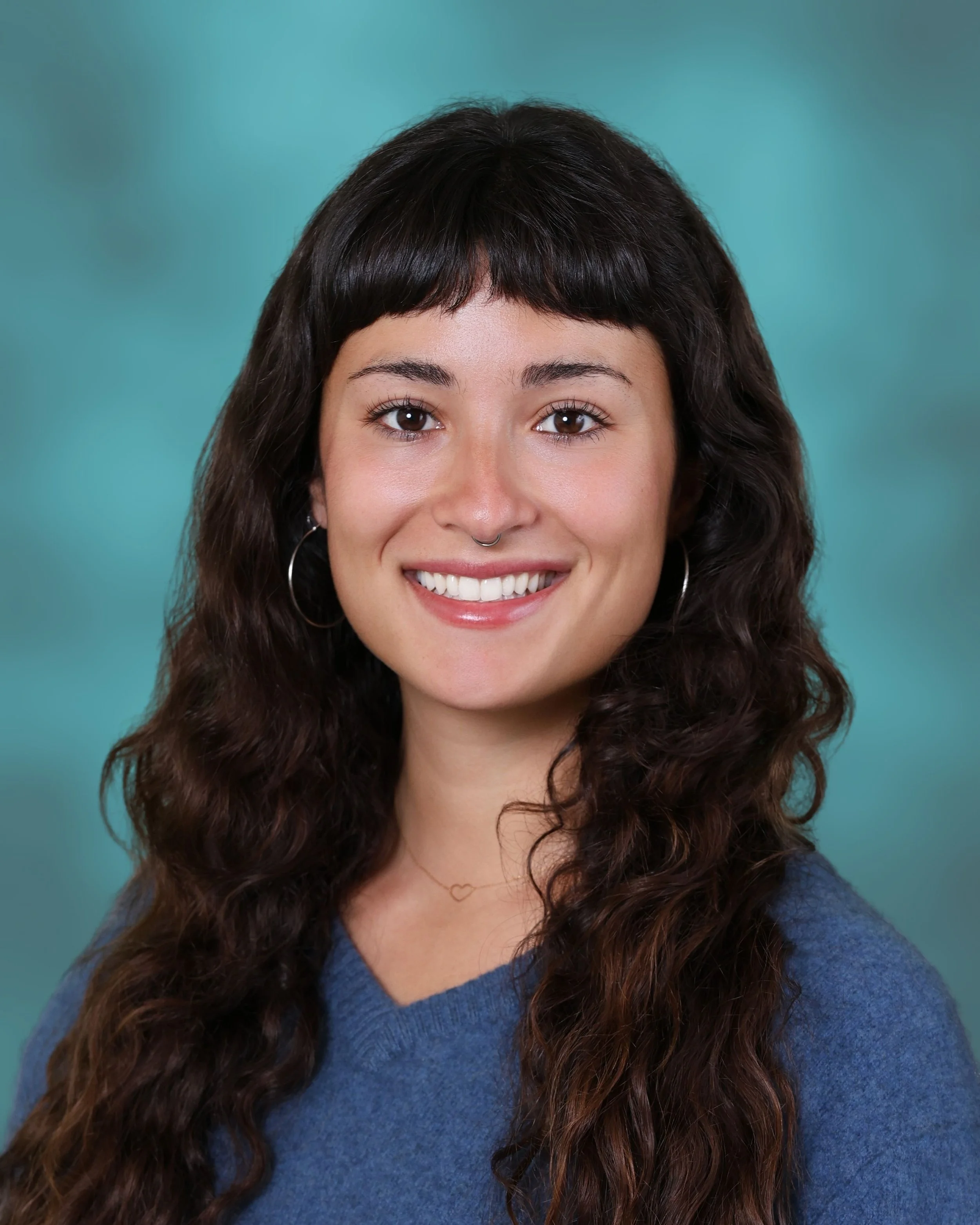 Close-up portrait of a woman with long curly brown hair, light skin, smiling showing white teeth, wearing a blue top, silver hoop earrings, and a heart-shaped necklace, with a teal background.