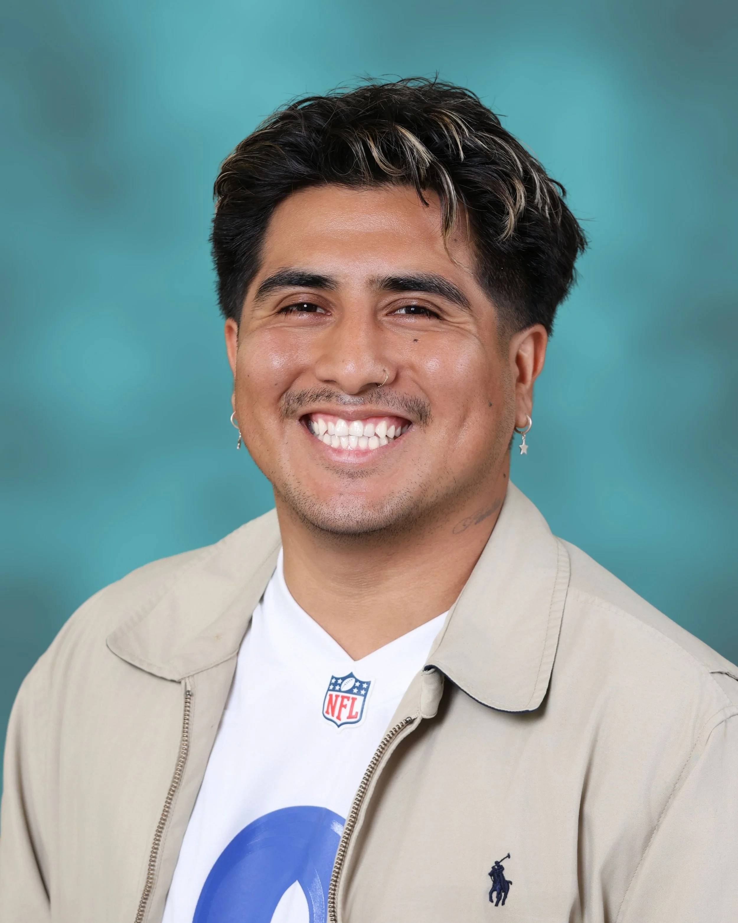 Close-up of a smiling man with dark hair, wearing a beige jacket and a white shirt with a partial blue logo, against a teal background.