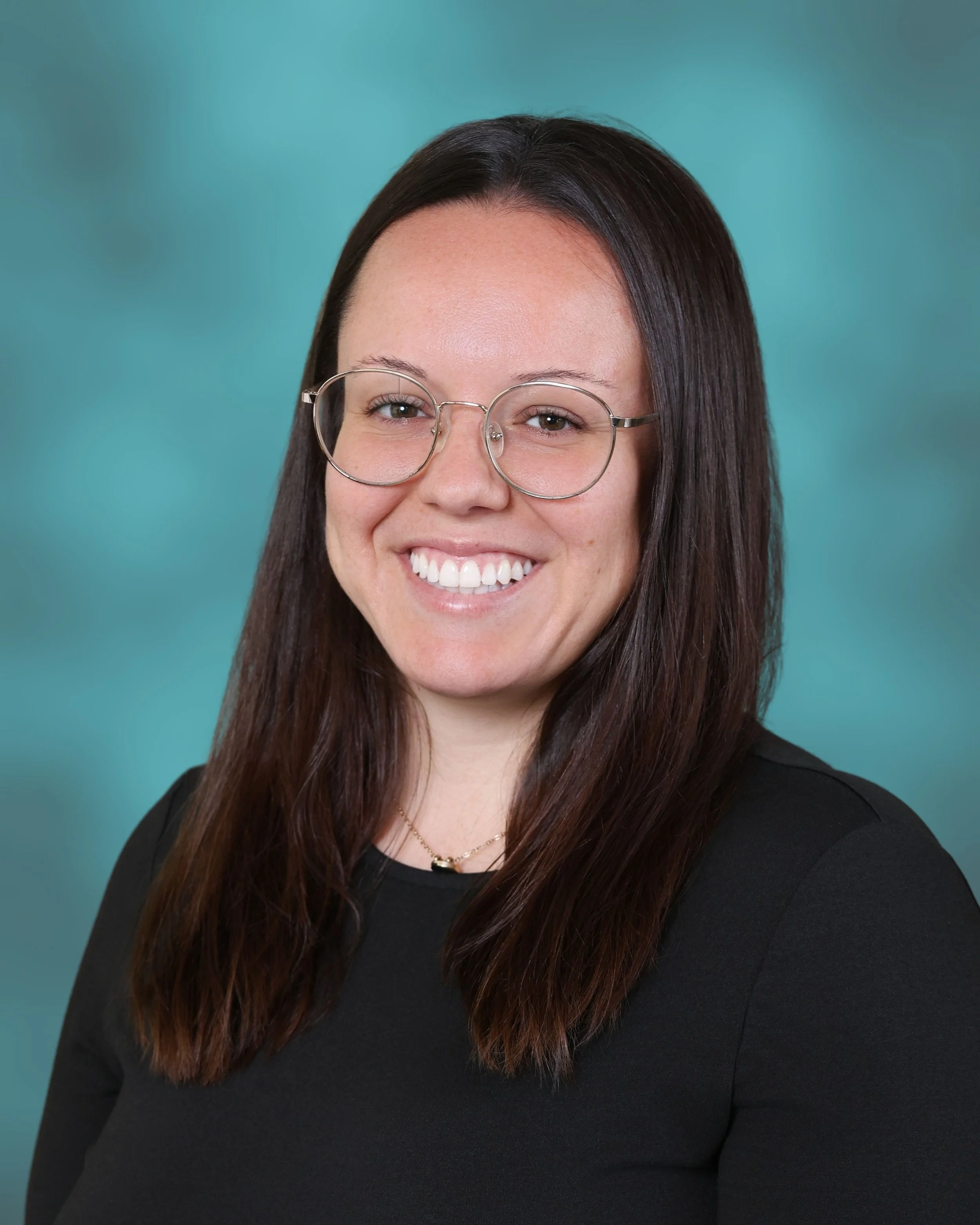 Smiling woman with long dark hair, wearing glasses and a black top, against a teal background.