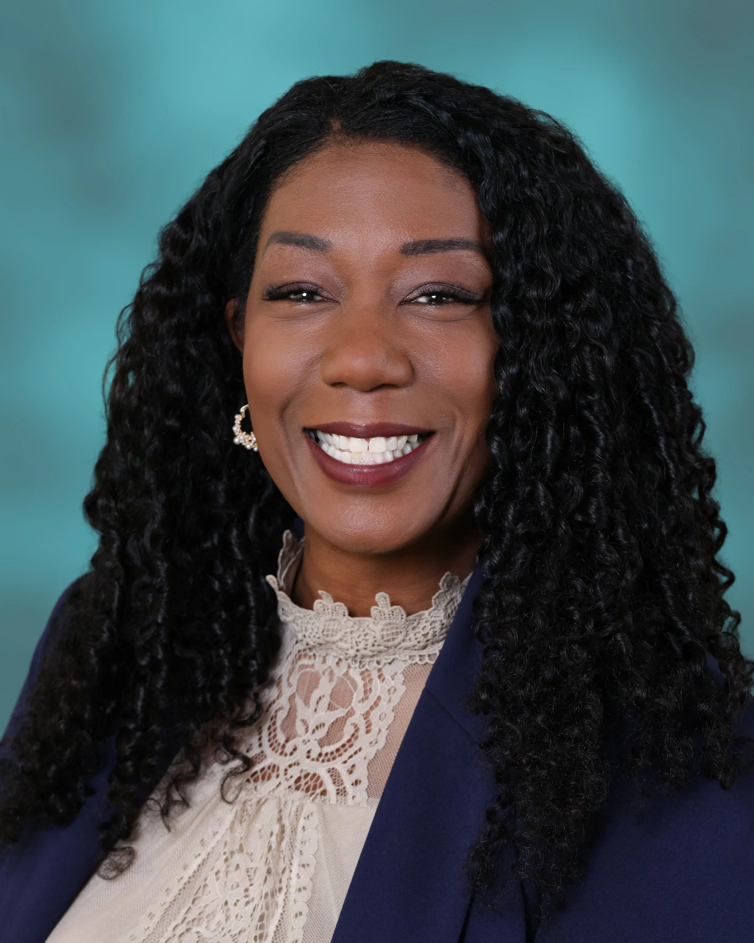 Close-up of a smiling woman with curly black hair, wearing a navy blazer and a beige lace top, against a teal background.