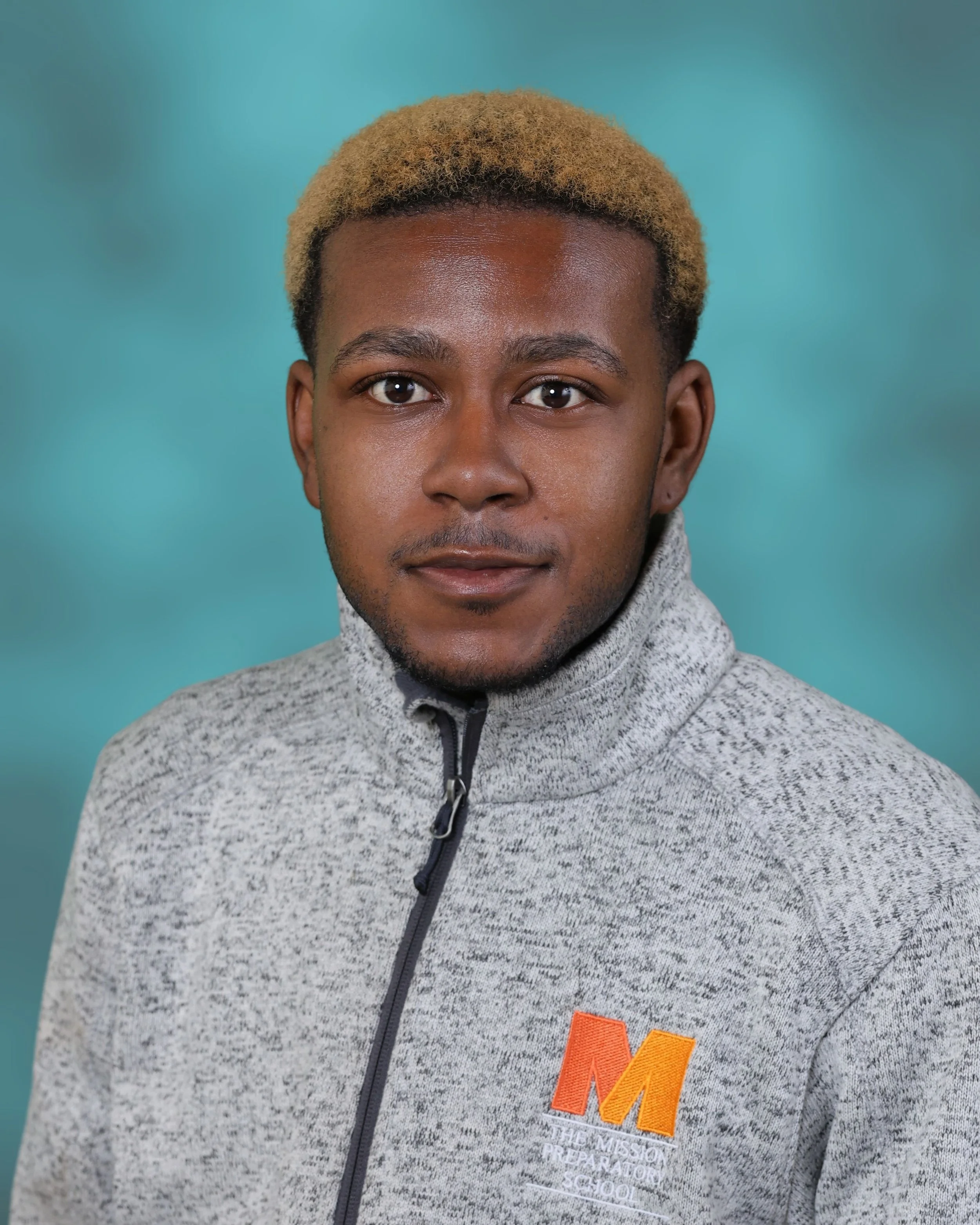 Close-up of a man with short, blonde hair, wearing a grey jacket with a zipper, against a blue-green background.