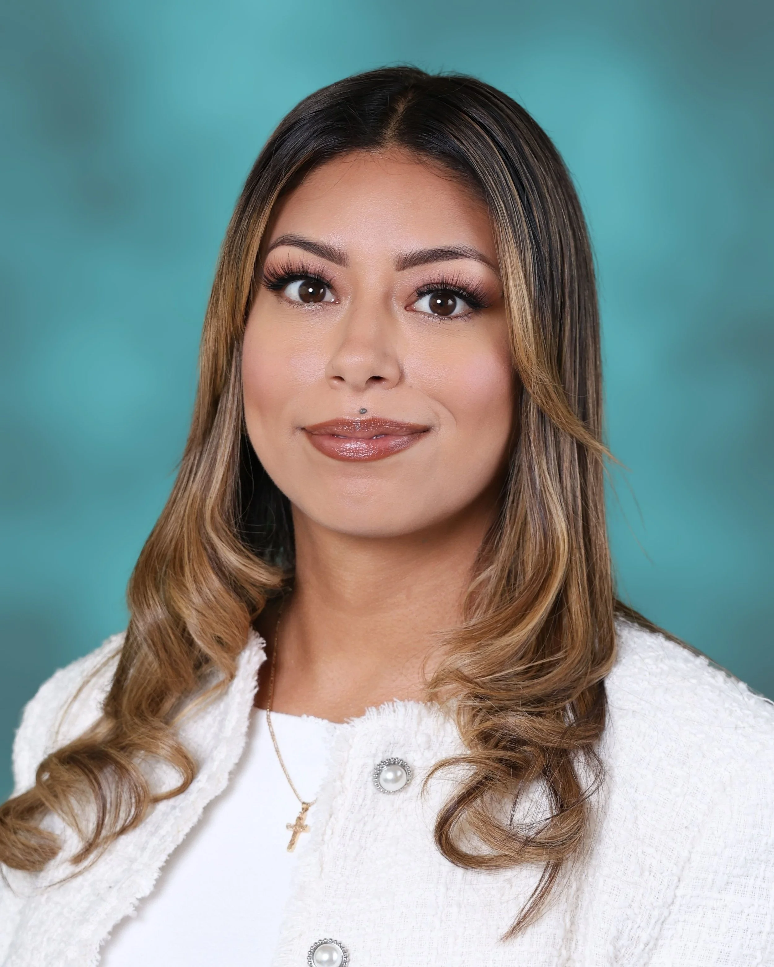 Close-up of a woman with styled brown hair with blonde highlights, wearing makeup with defined eyebrows, long eyelashes, and glossy lips, dressed in a white textured cardigan with pearl and gold jewelry, against a teal background.