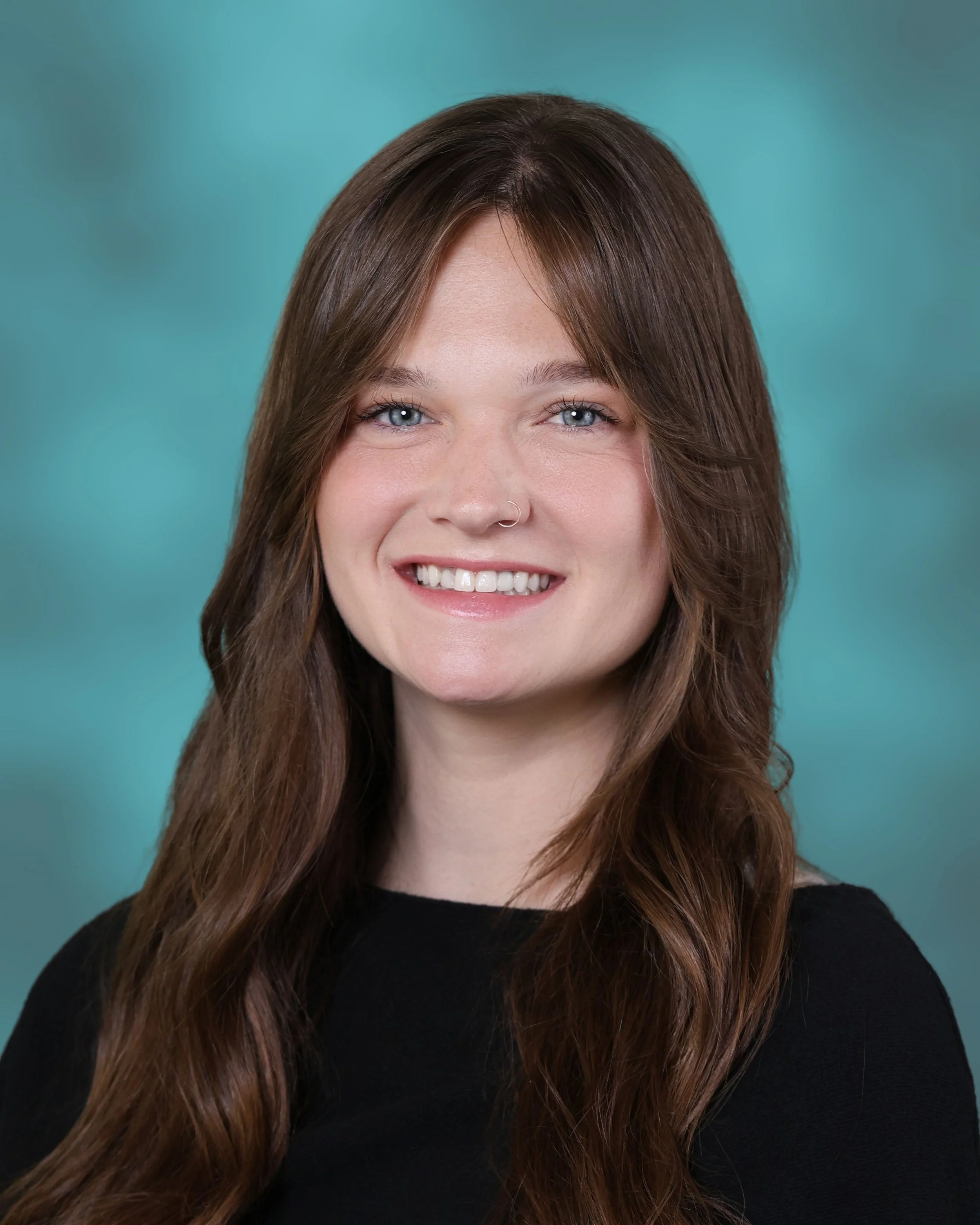 Headshot of a smiling woman with shoulder-length brown hair and blue eyes, wearing a black top, against a teal background.