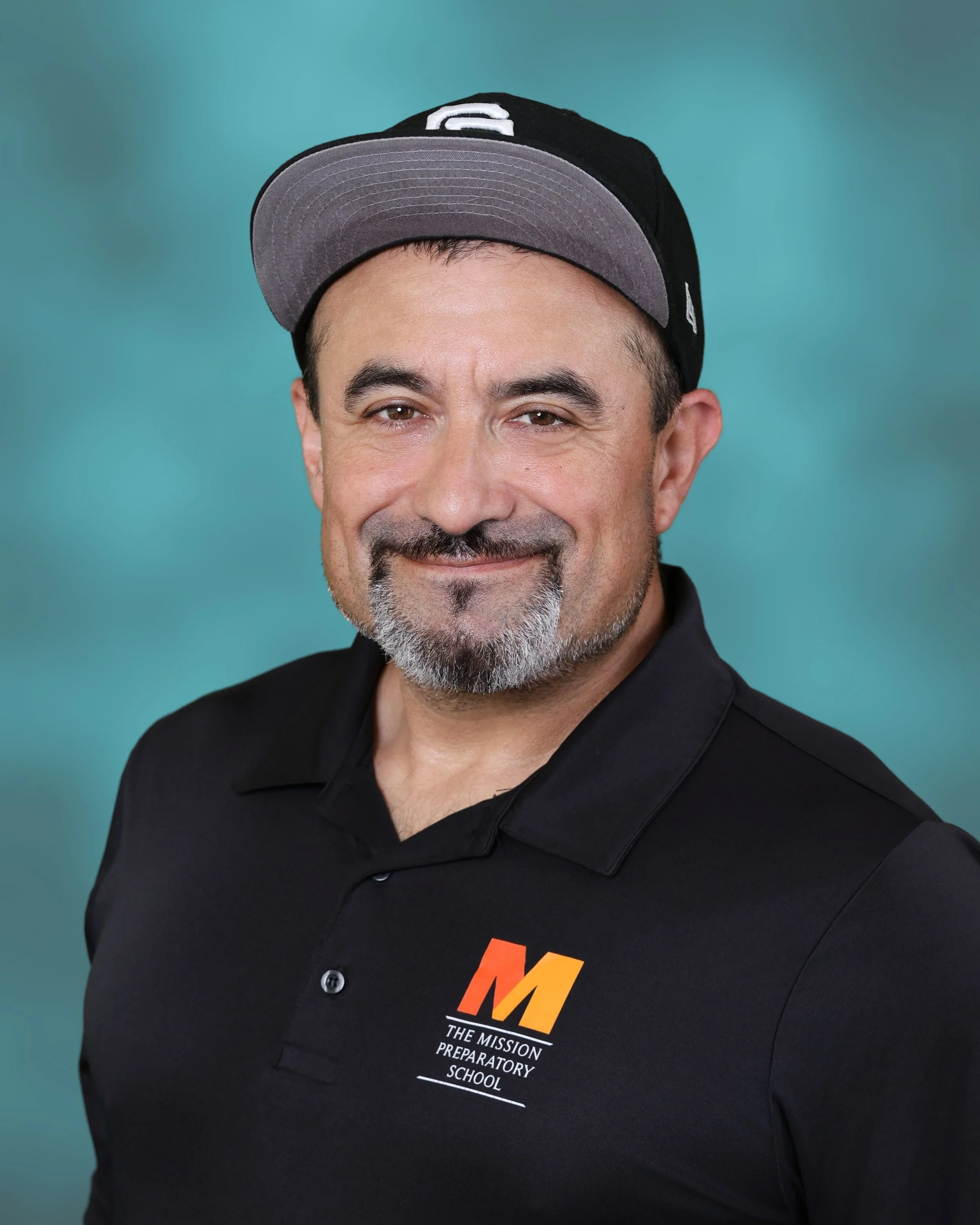A man with a beard wearing a black cap and black polo shirt with a logo that reads 'The Mission Preparatory School' in orange, white, and gray, smiling against a turquoise background.