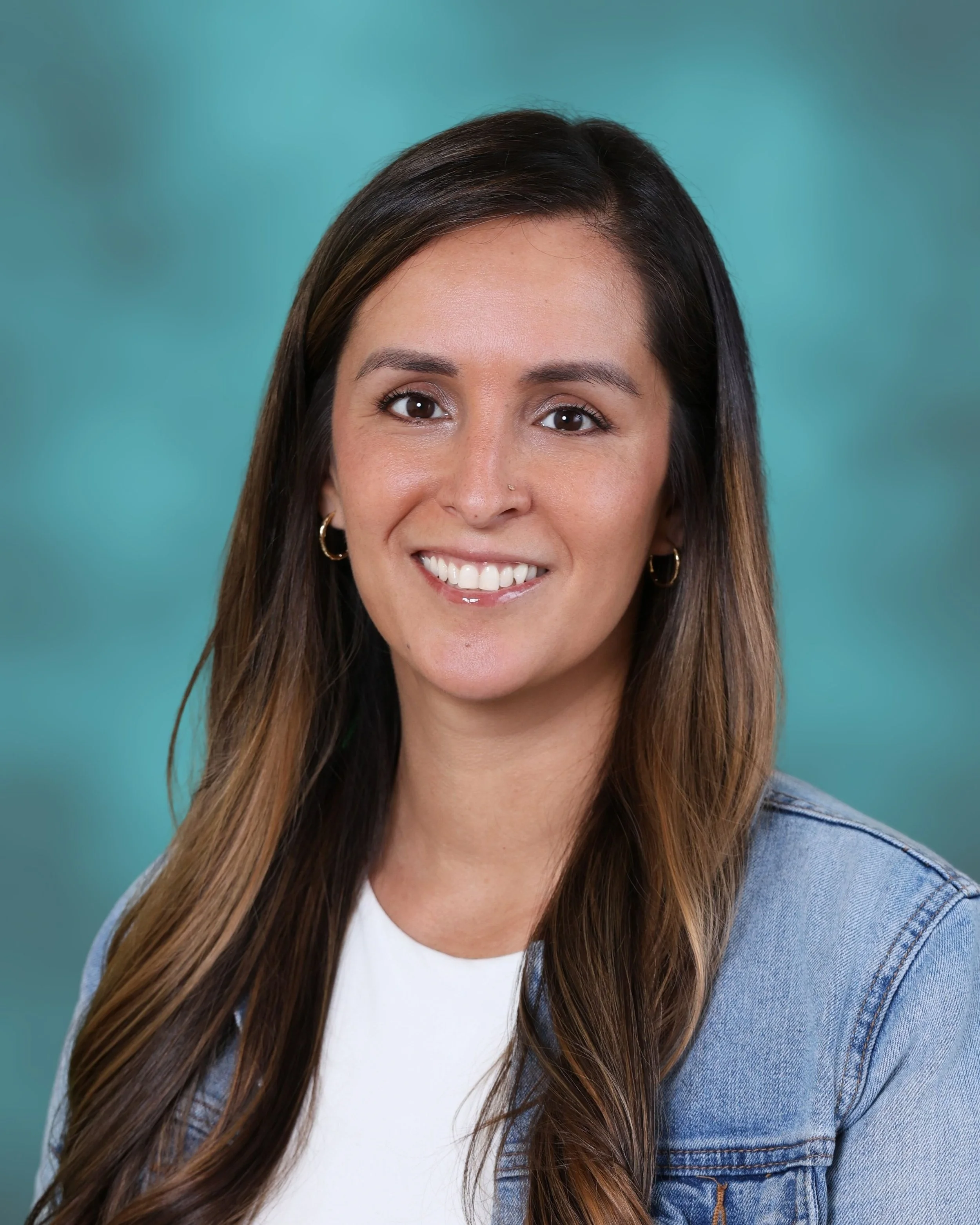 A woman with long brown hair, wearing hoop earrings, smiling, a white shirt, and a denim jacket, against a teal background.