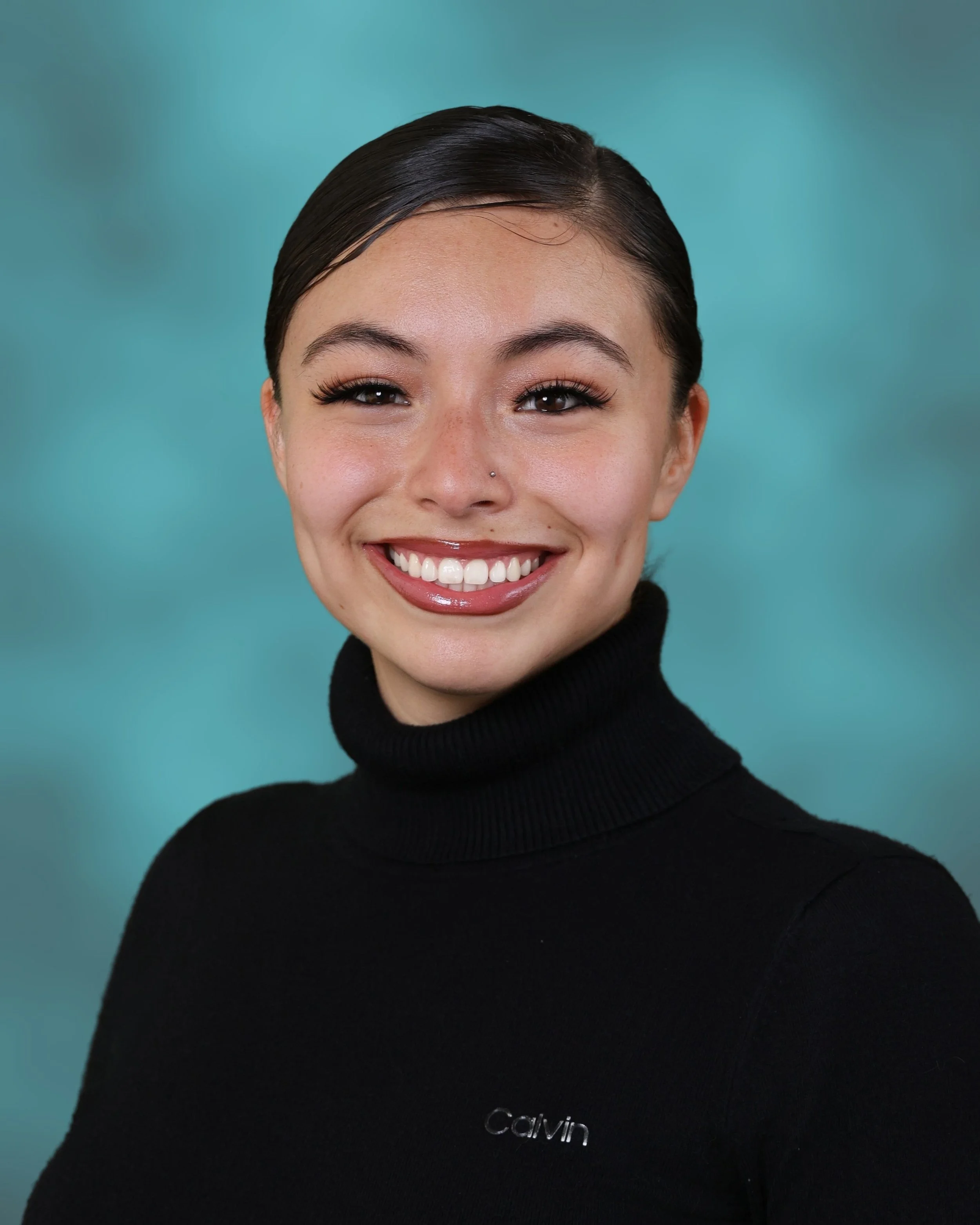 Close-up of a smiling woman with dark hair slicked back, wearing a black Calvin Klein turtleneck sweater, against a blue-green gradient background.
