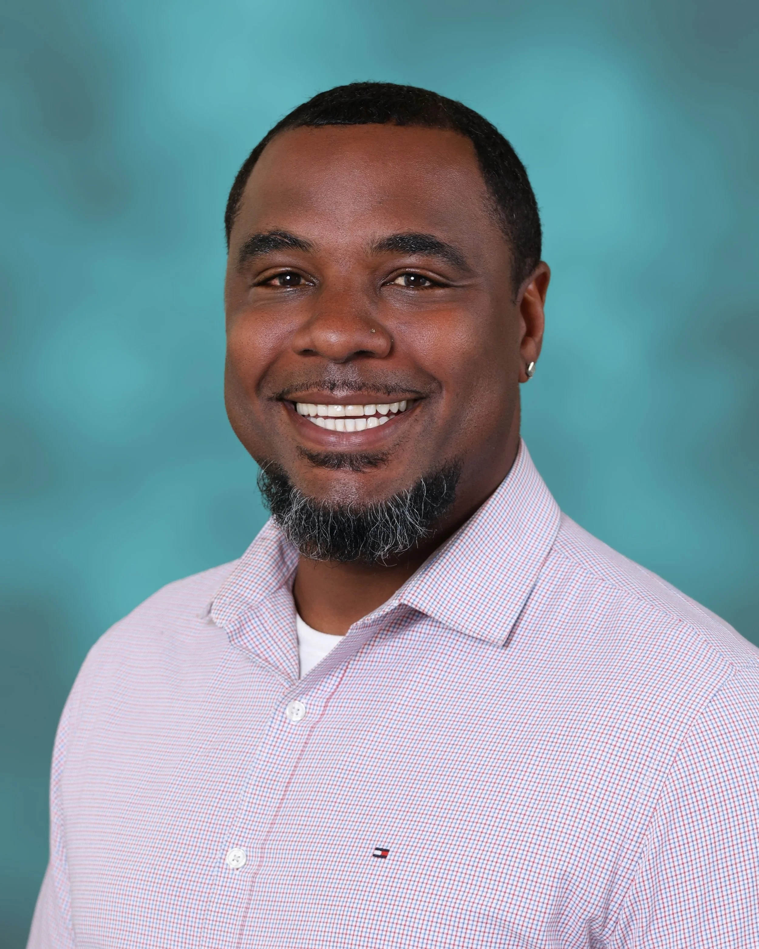 Portrait of a man with short black hair and a beard, smiling, wearing a pink checkered shirt against a teal background.