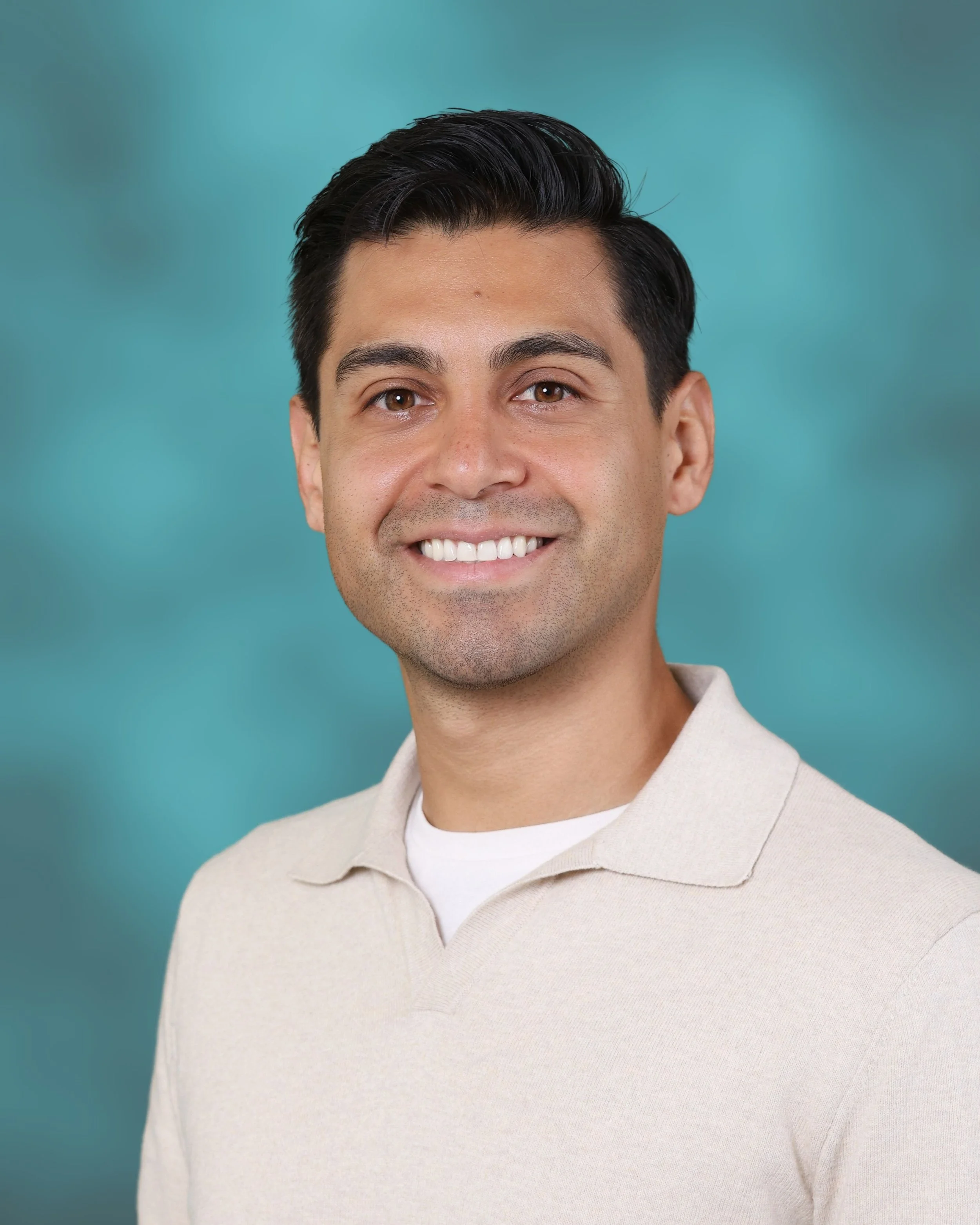 Close-up of a smiling man with short dark hair, wearing a beige collar shirt, with a plain teal background.