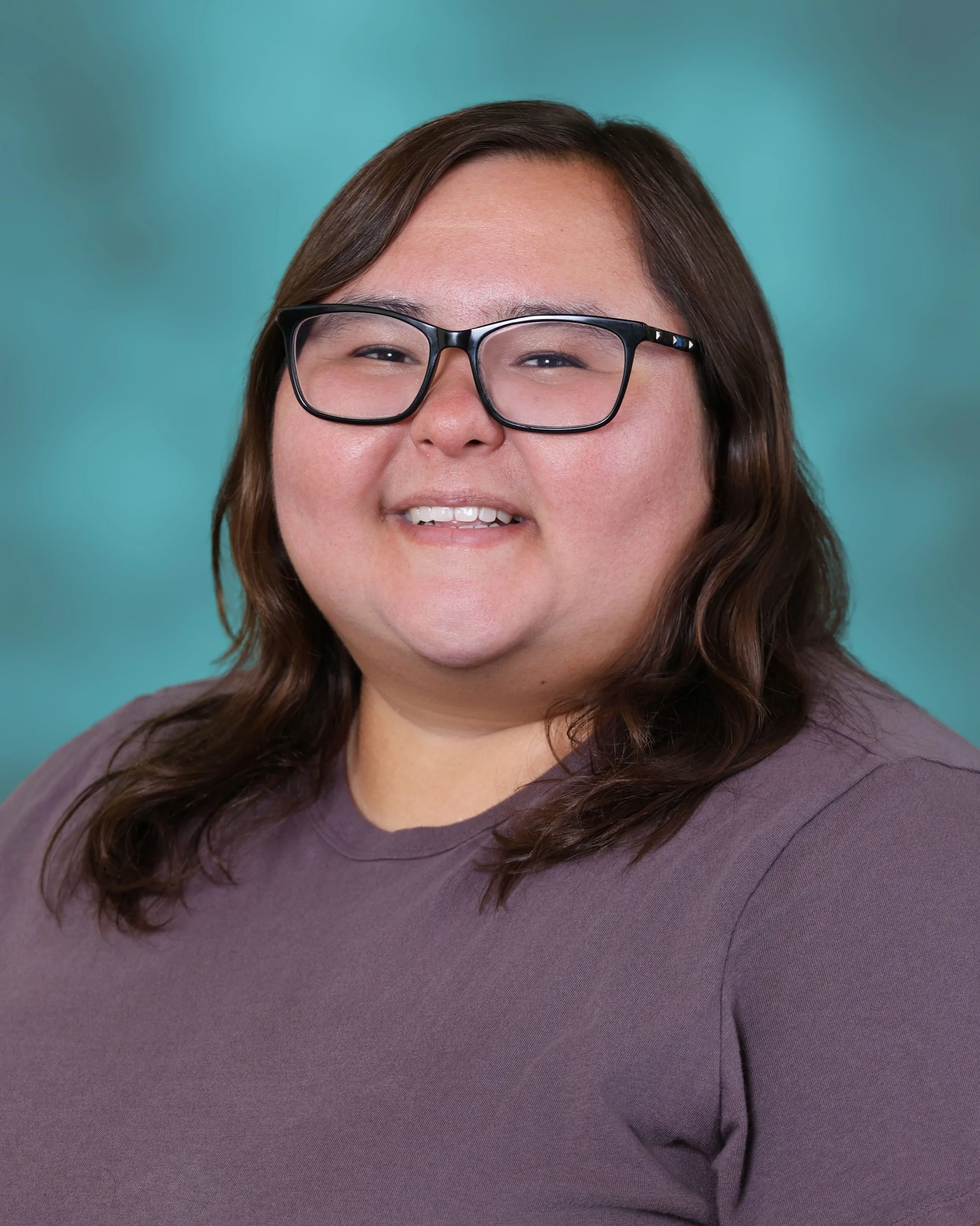 Close-up of a smiling woman with glasses, brown hair, wearing a purple top, against a teal background.