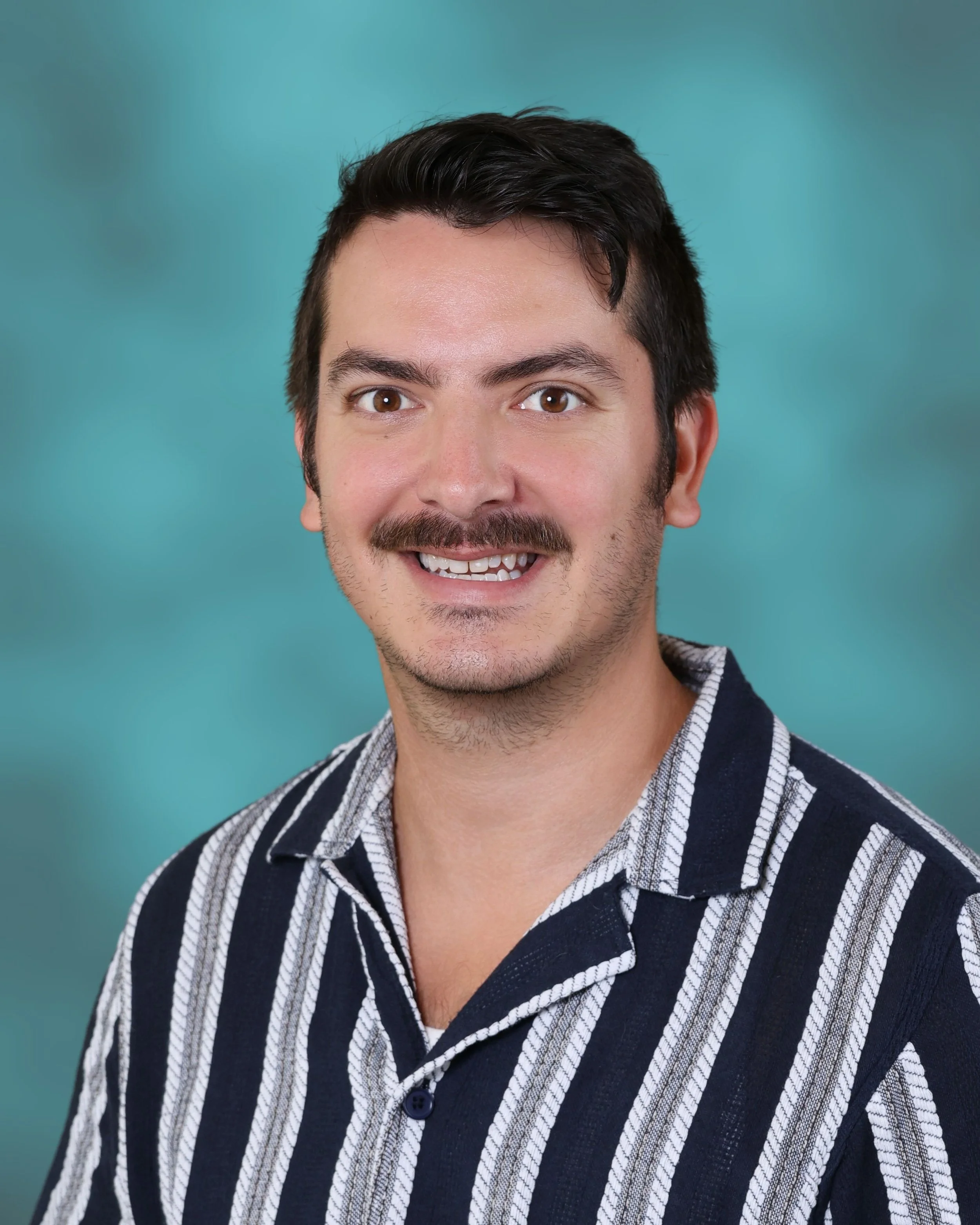 Close-up of a smiling man with dark hair, facial hair, wearing a striped shirt, against a teal background.