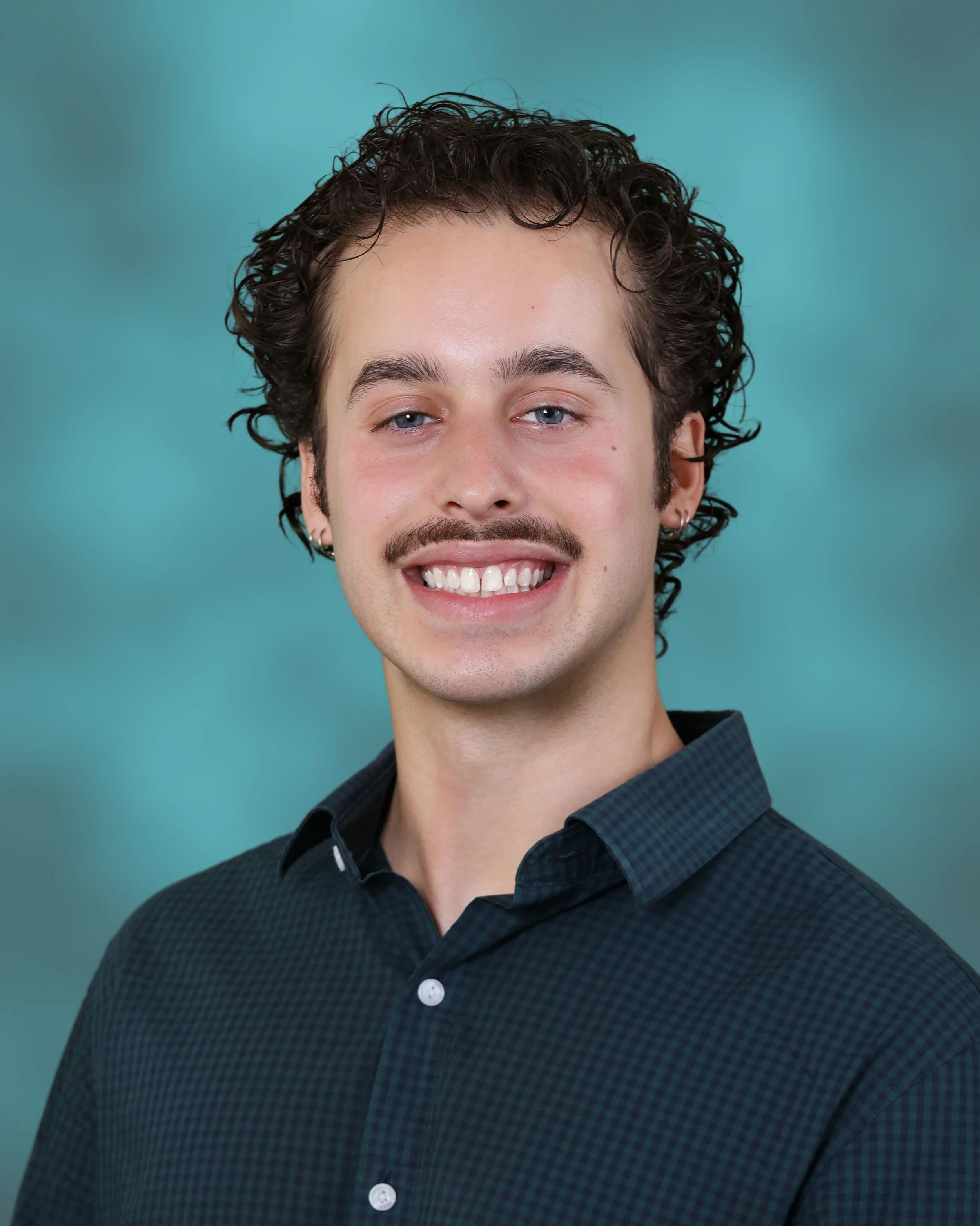 Young man with curly hair, blue eyes, and a beard, smiling in front of a teal background.