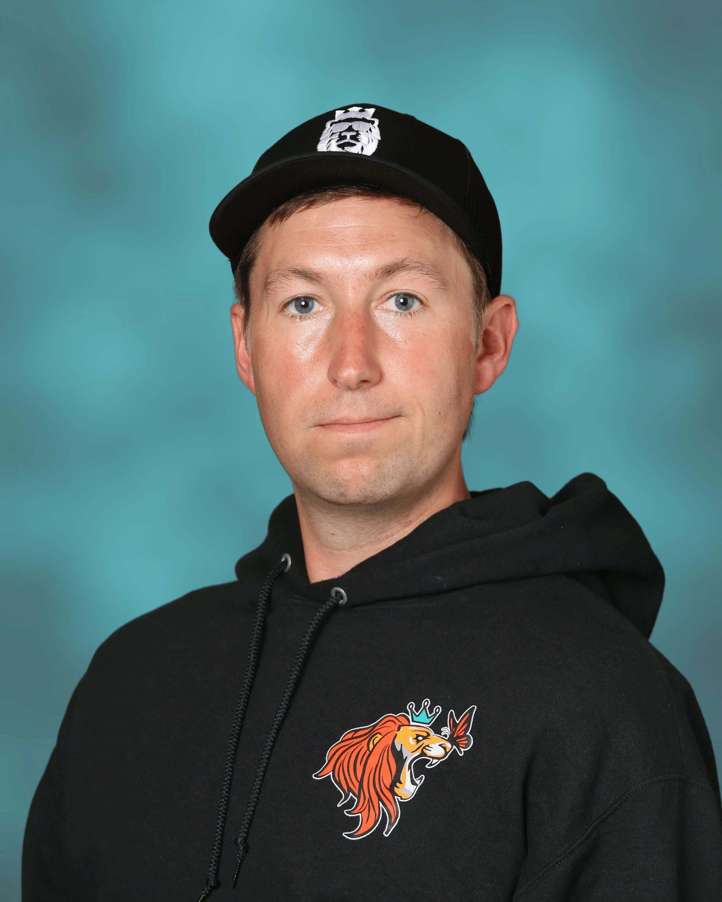 A young man with light skin, blue eyes, and short brown hair wearing a black baseball cap with a white lion head logo and a black hoodie with a colorful lion graphic.