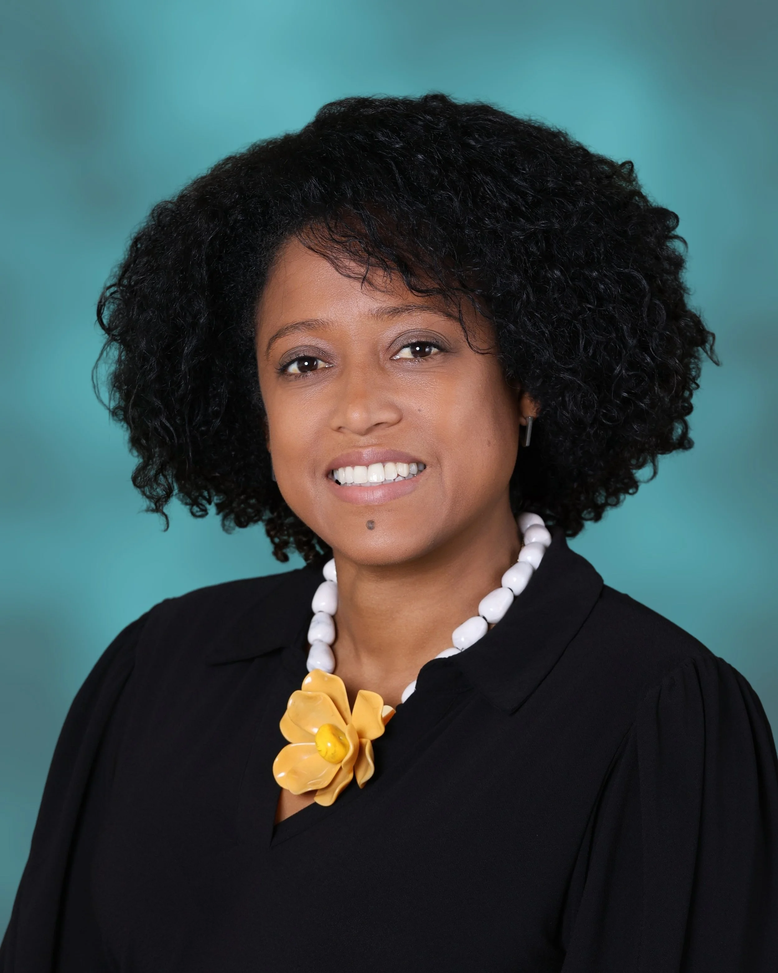 Close-up portrait of a smiling woman with curly black hair, wearing a black top, a white beaded necklace, and a large yellow flower necklace, against a teal background.