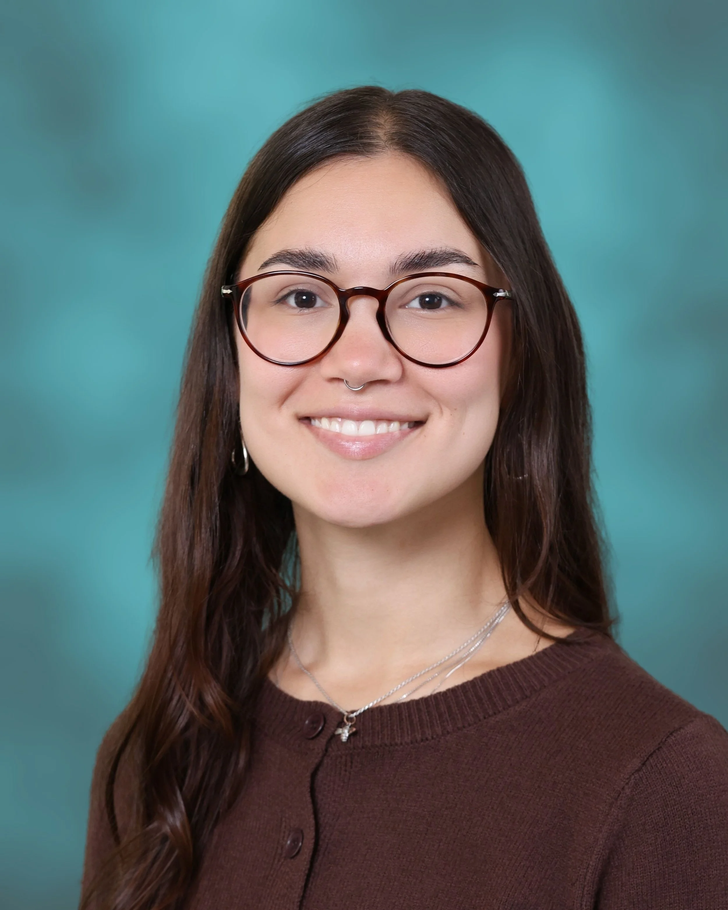 Close-up portrait of a young woman with long brown hair, wearing glasses, a brown sweater, and silver jewelry, smiling against a teal background.