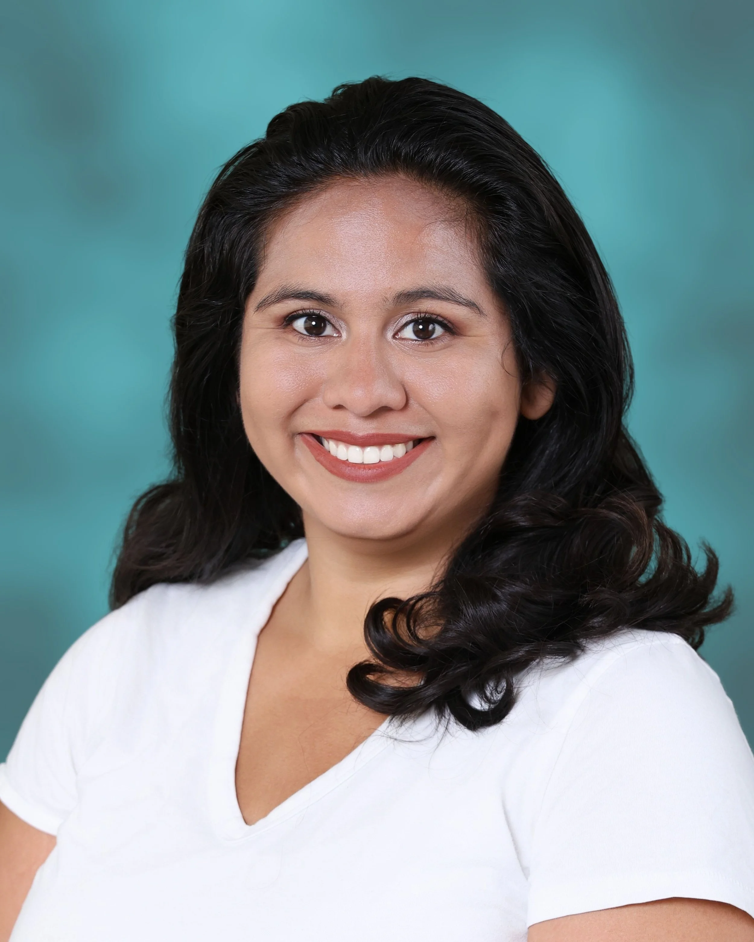 Close-up portrait of a smiling woman with dark, wavy hair wearing a white top with a V-neckline against a teal background.