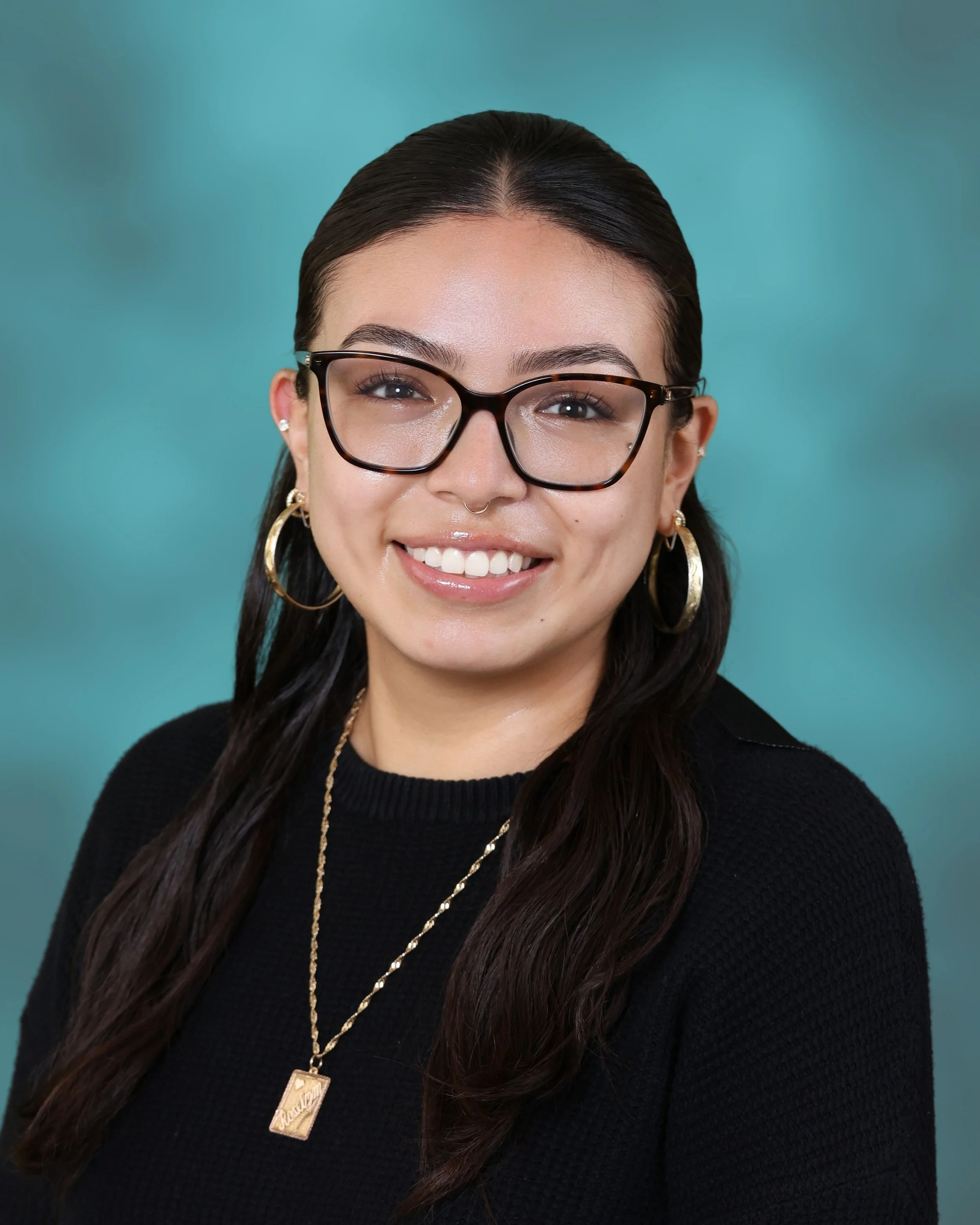 Close-up portrait of a young woman with dark hair, wearing glasses, gold hoop earrings, and a black sweater, smiling, with a teal background.