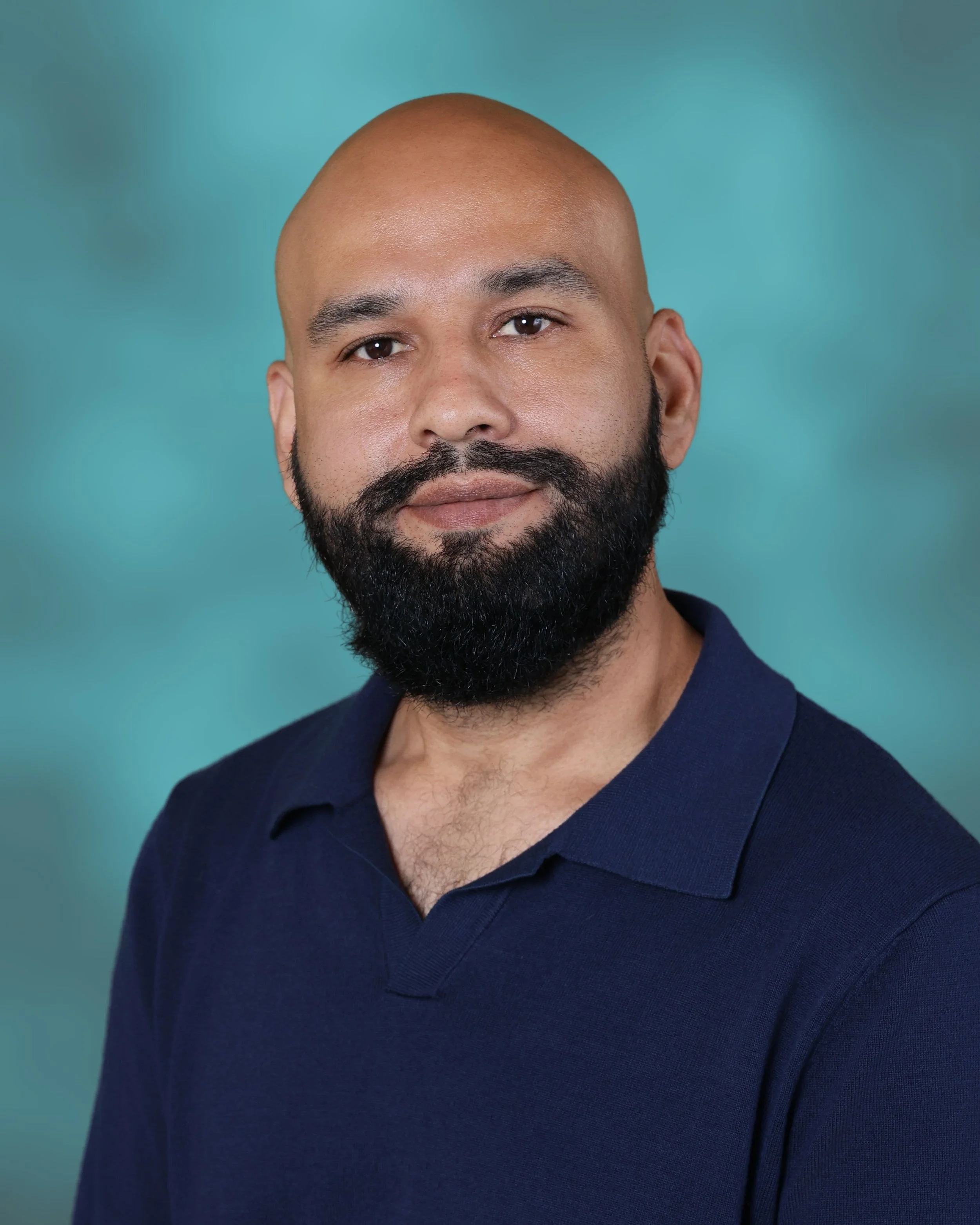 A man with a neatly groomed beard, wearing a dark blue collared shirt, smiling gently against a teal background.