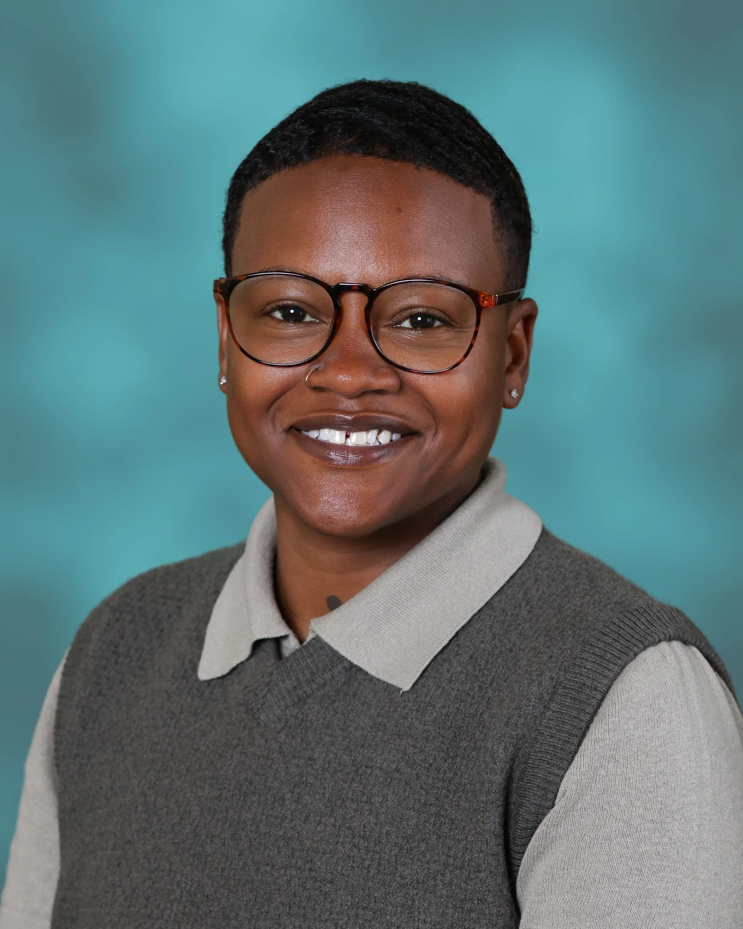 A young woman with dark skin, short black hair, glasses, and pearl earrings, smiling in front of a teal background, wearing a gray collared shirt under a gray sweater.