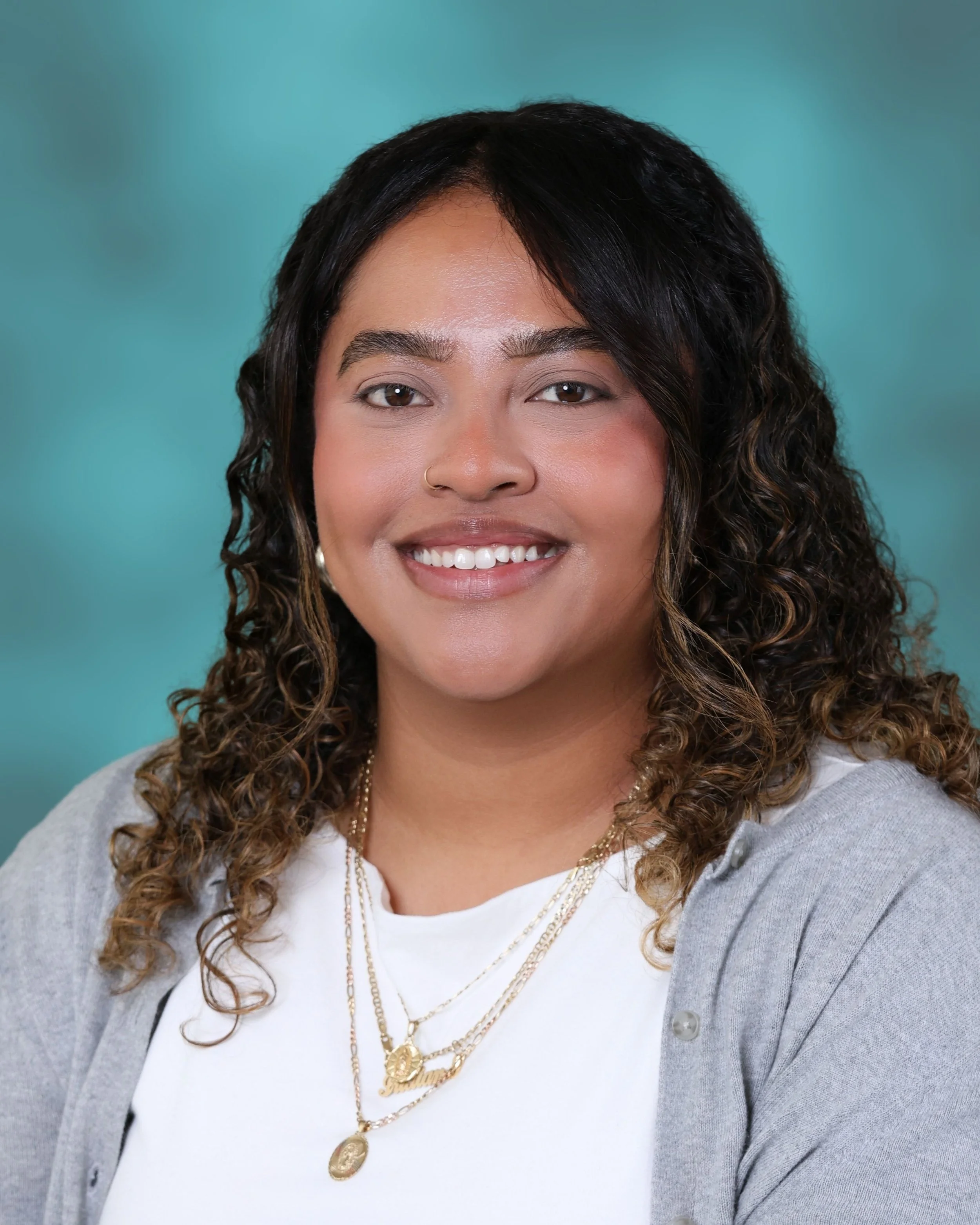 Close-up of a smiling woman with curly hair, wearing a gray cardigan, a white t-shirt, and layered gold necklaces, against a teal background.