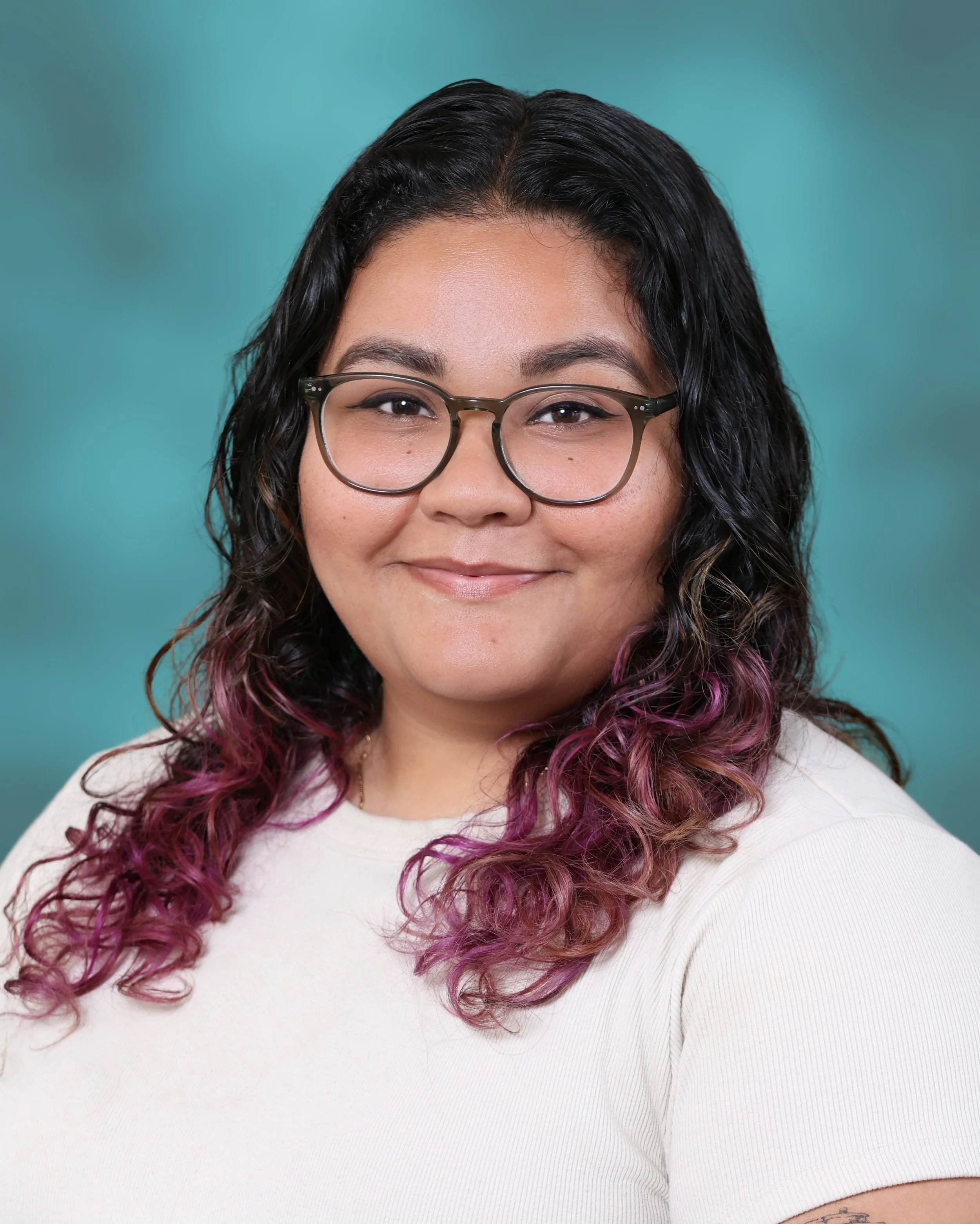 Close-up portrait of a smiling woman with curly black and purple hair, wearing glasses and a light-colored sweater, against a teal background.