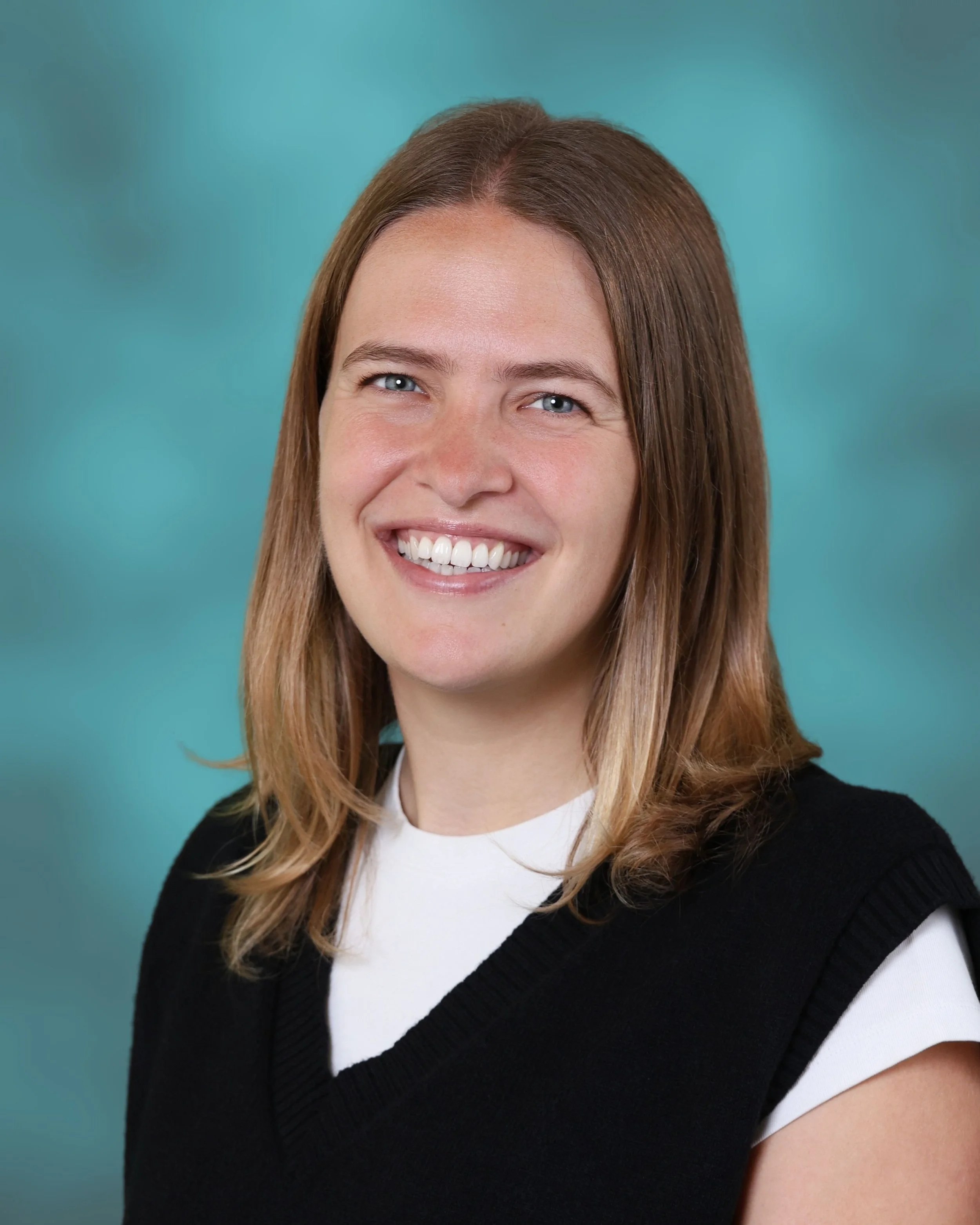A woman with long, light brown hair smiling, wearing a black sweater over a white shirt, against a teal background.