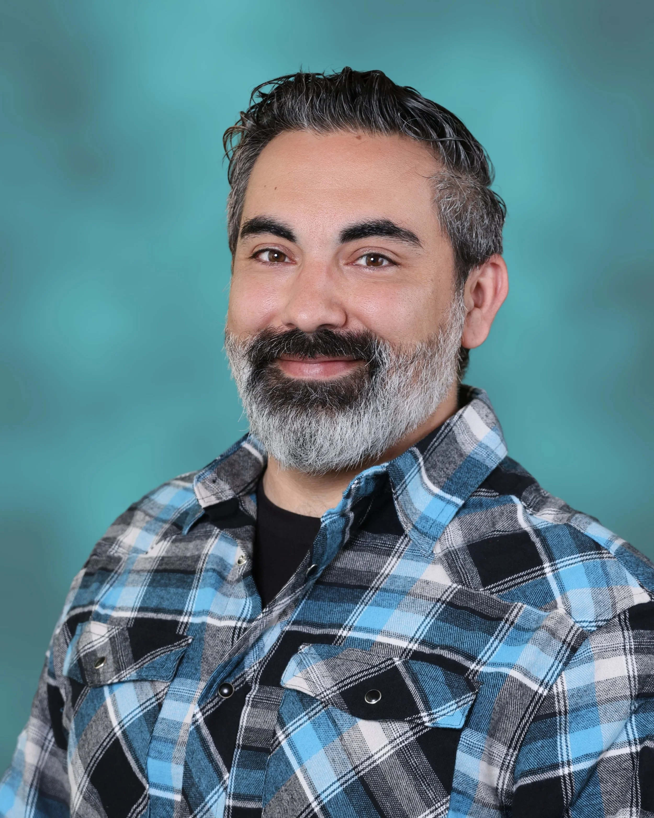 A close-up of a middle-aged man with dark hair and a salt-and-pepper beard, wearing a blue and black plaid shirt, smiling softly, with a blurred teal background.