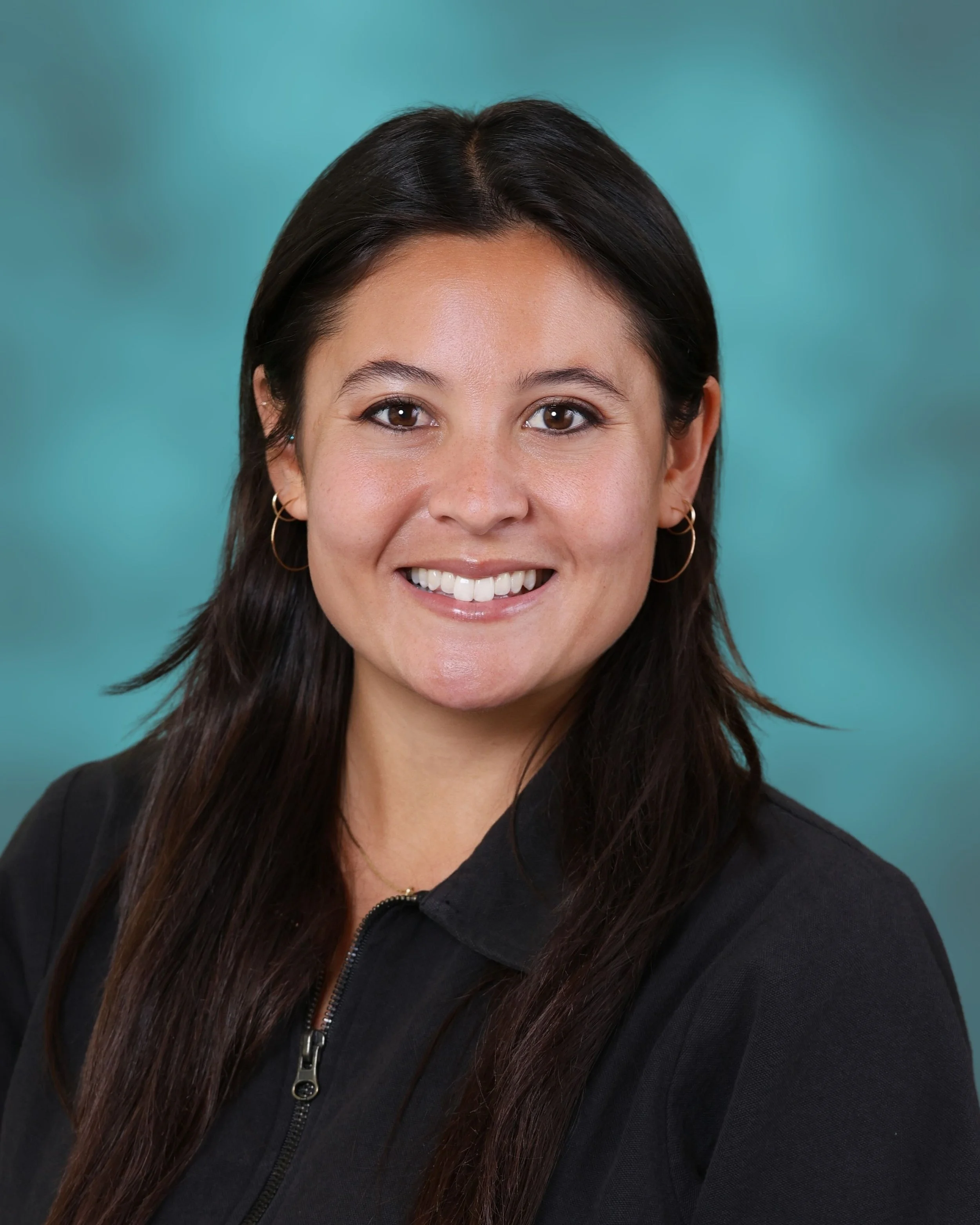 Close-up portrait of a woman with dark hair, brown eyes, and gold hoop earrings smiling, wearing a black zip-up jacket, against a teal background.