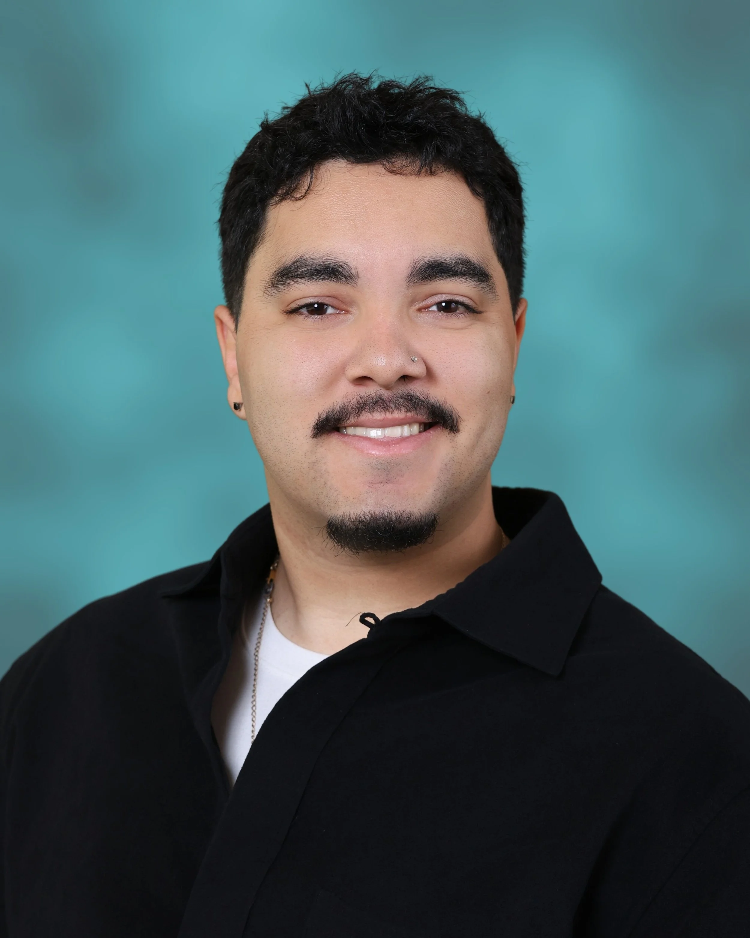 A young man with dark, curly hair and a beard, smiling and wearing a black jacket over a white shirt, against a teal background.
