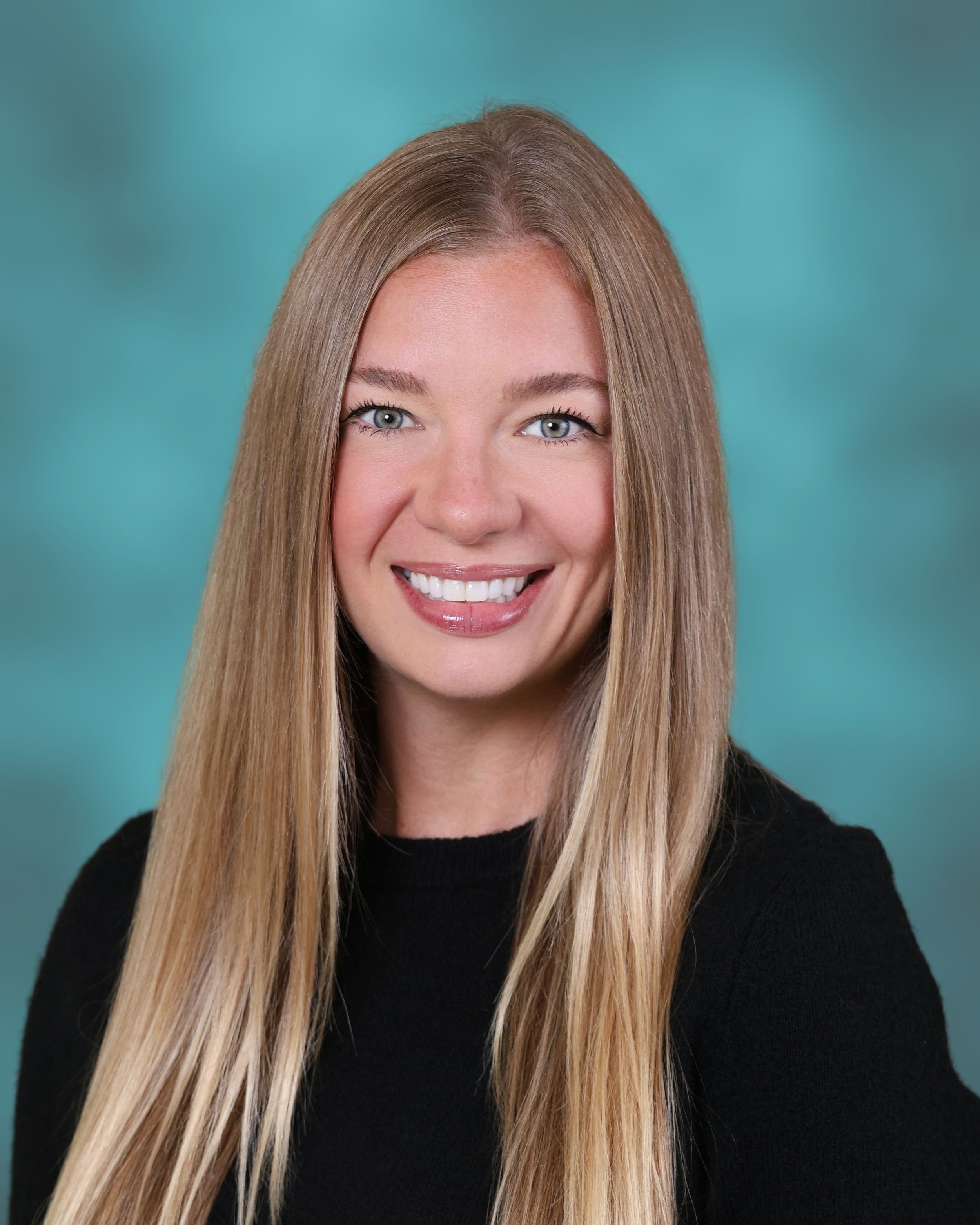 Close-up of a smiling woman with long blonde hair, pink lipstick, and wearing a black sweater, against a teal background.