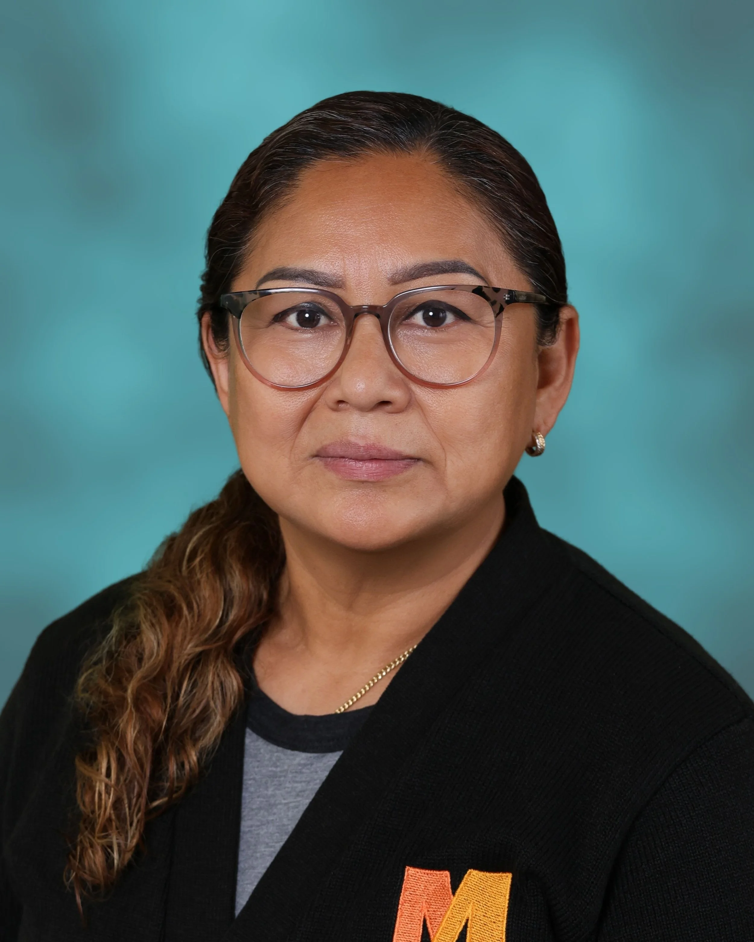 Close-up of a woman with dark hair tied back, wearing glasses and a black university sweatshirt with an orange and yellow letter 'M' logo, against a teal background.