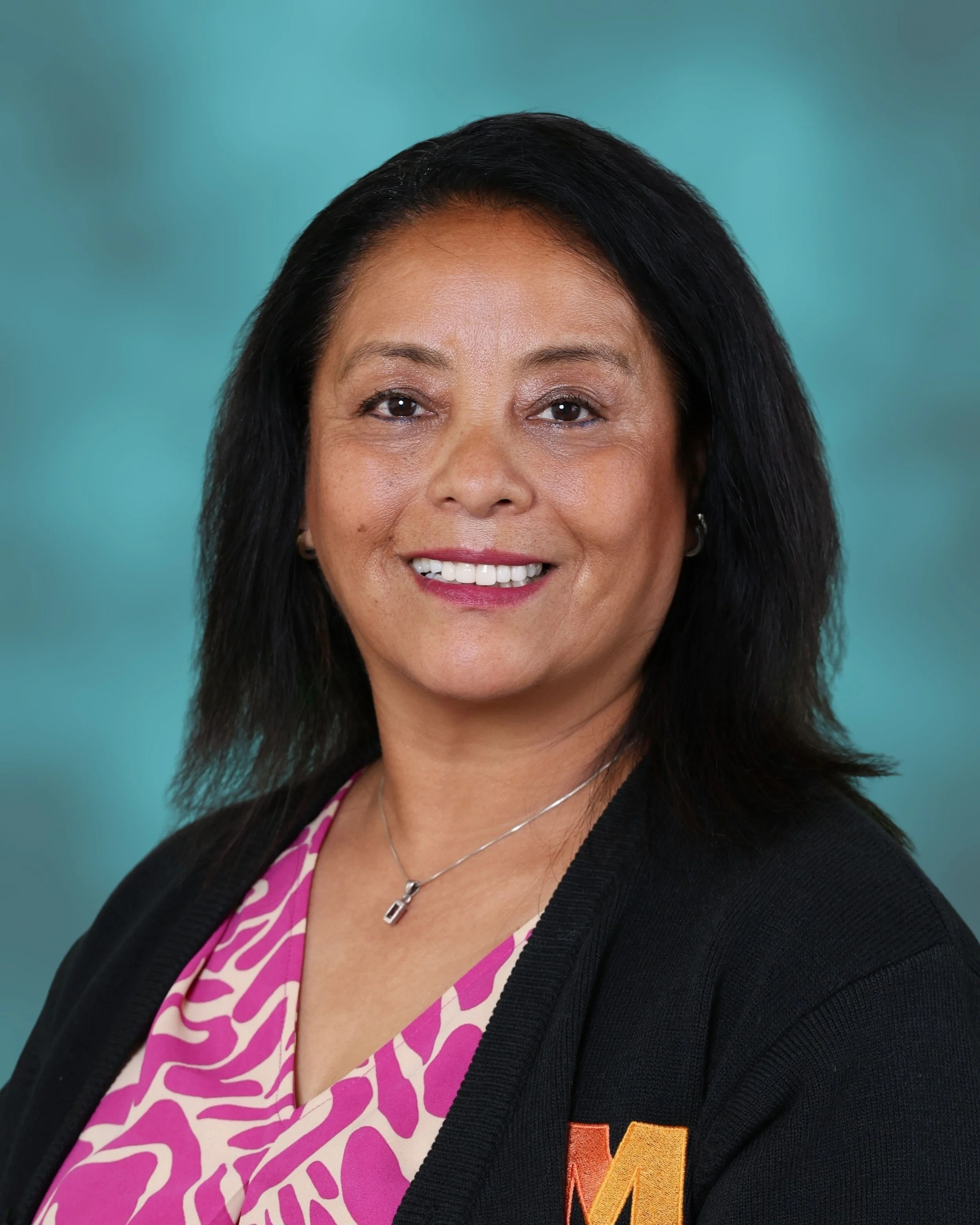 A woman with black hair smiling, wearing a pink and white patterned blouse, a black jacket, a necklace.