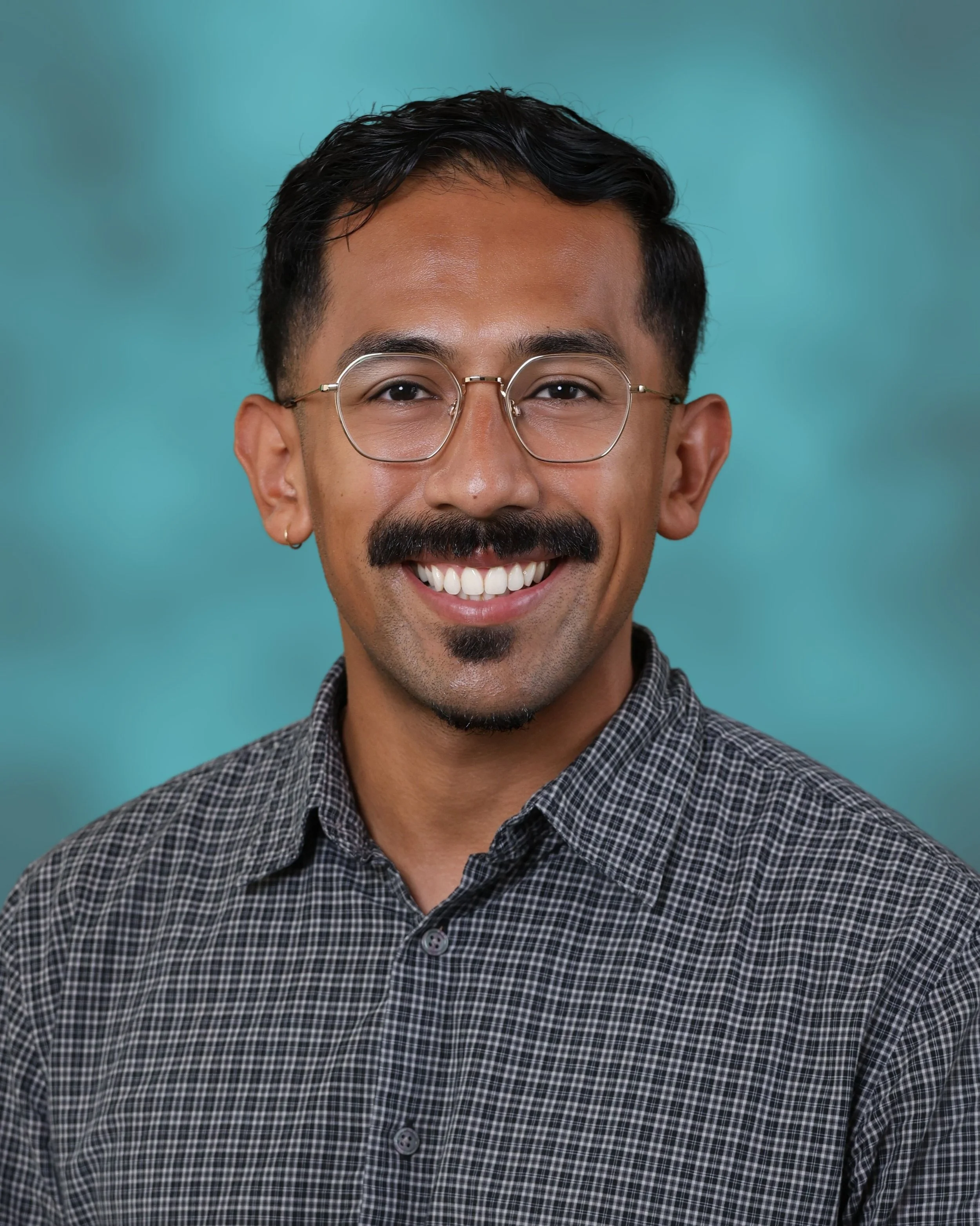 A smiling man with glasses, a mustache, a goatee, and hoop earrings, wearing a checkered shirt against a teal background.