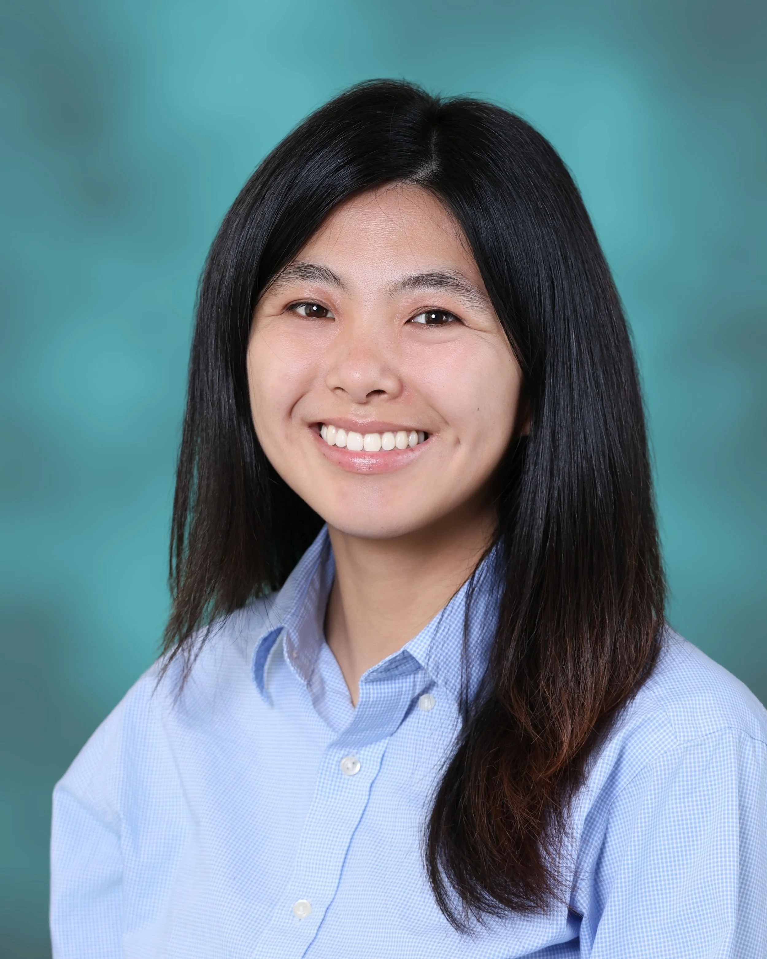 Close-up photo of a woman with long black hair, smiling, wearing a blue button-up shirt against a teal background.