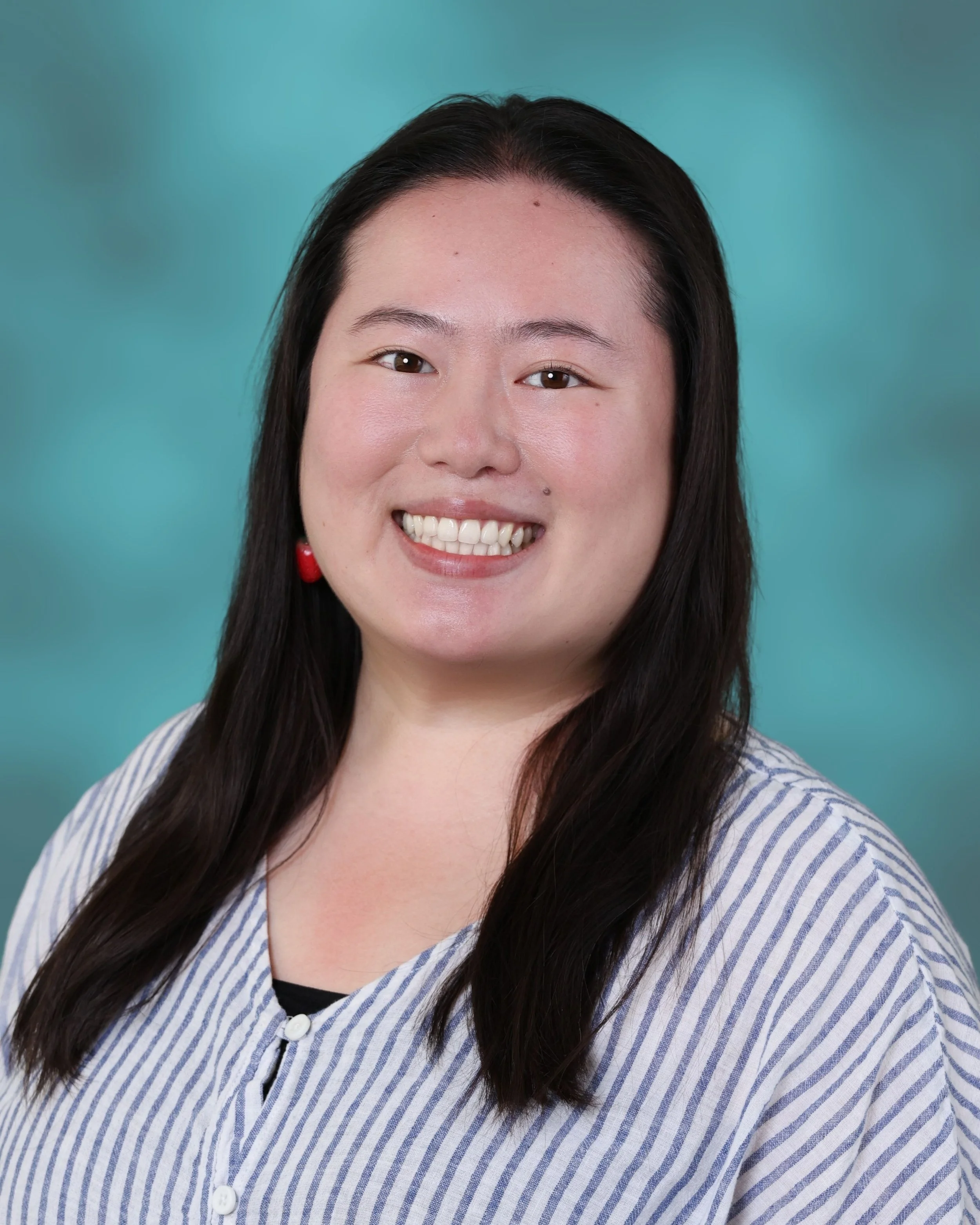 Close-up of a smiling woman with long dark hair, wearing a striped shirt and red earrings, against a teal background.