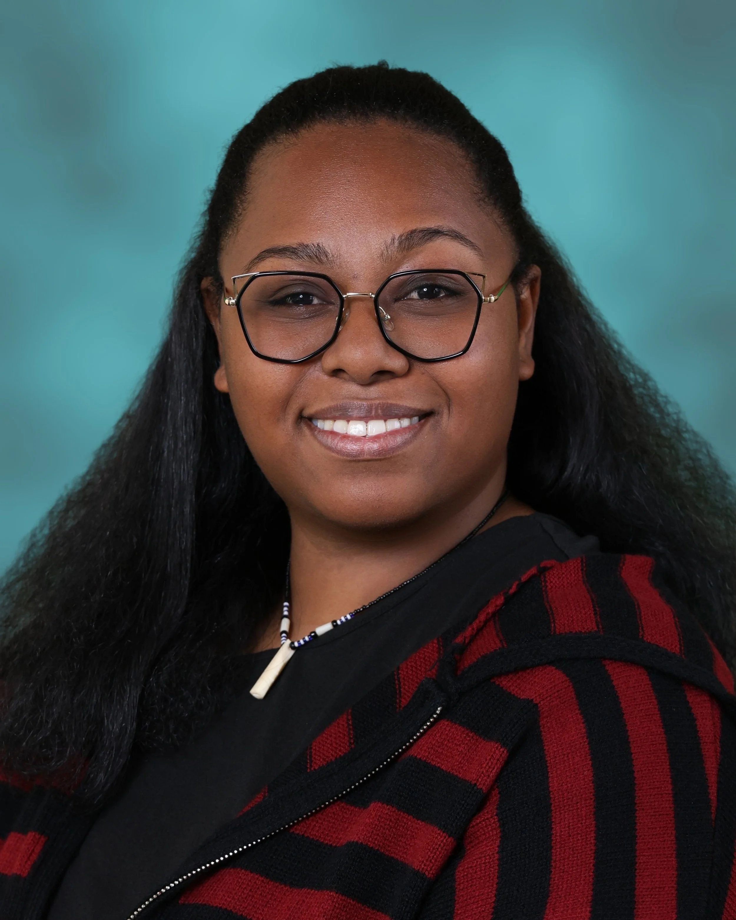 Close-up of a smiling woman with glasses, long dark hair, wearing a black and red patterned sweater, and a necklace with beads and a shell pendant, against a turquoise background.