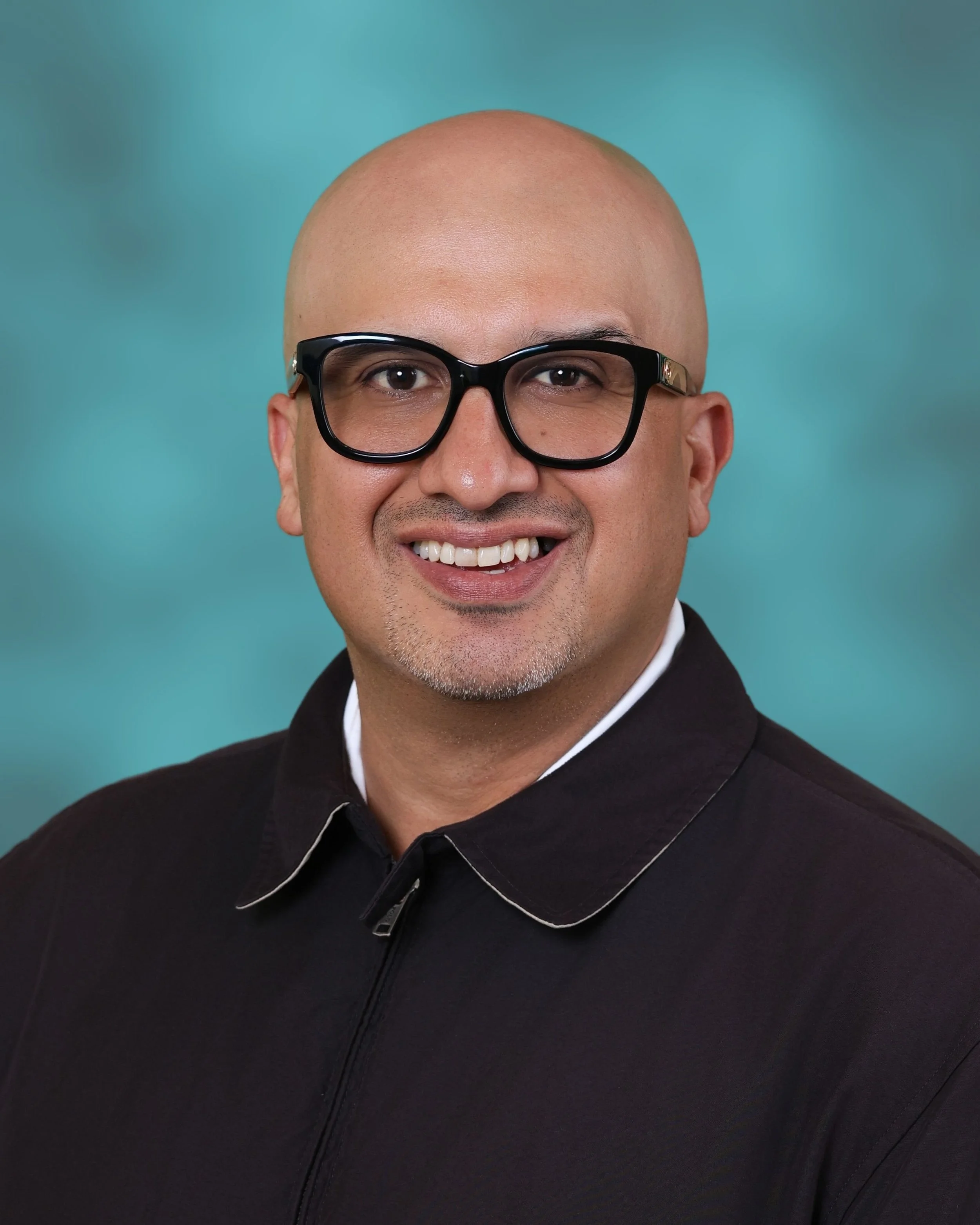 Man with a shaved head, black-rimmed glasses, a big smile showing teeth, wearing a black jacket with white trim, against a teal background.