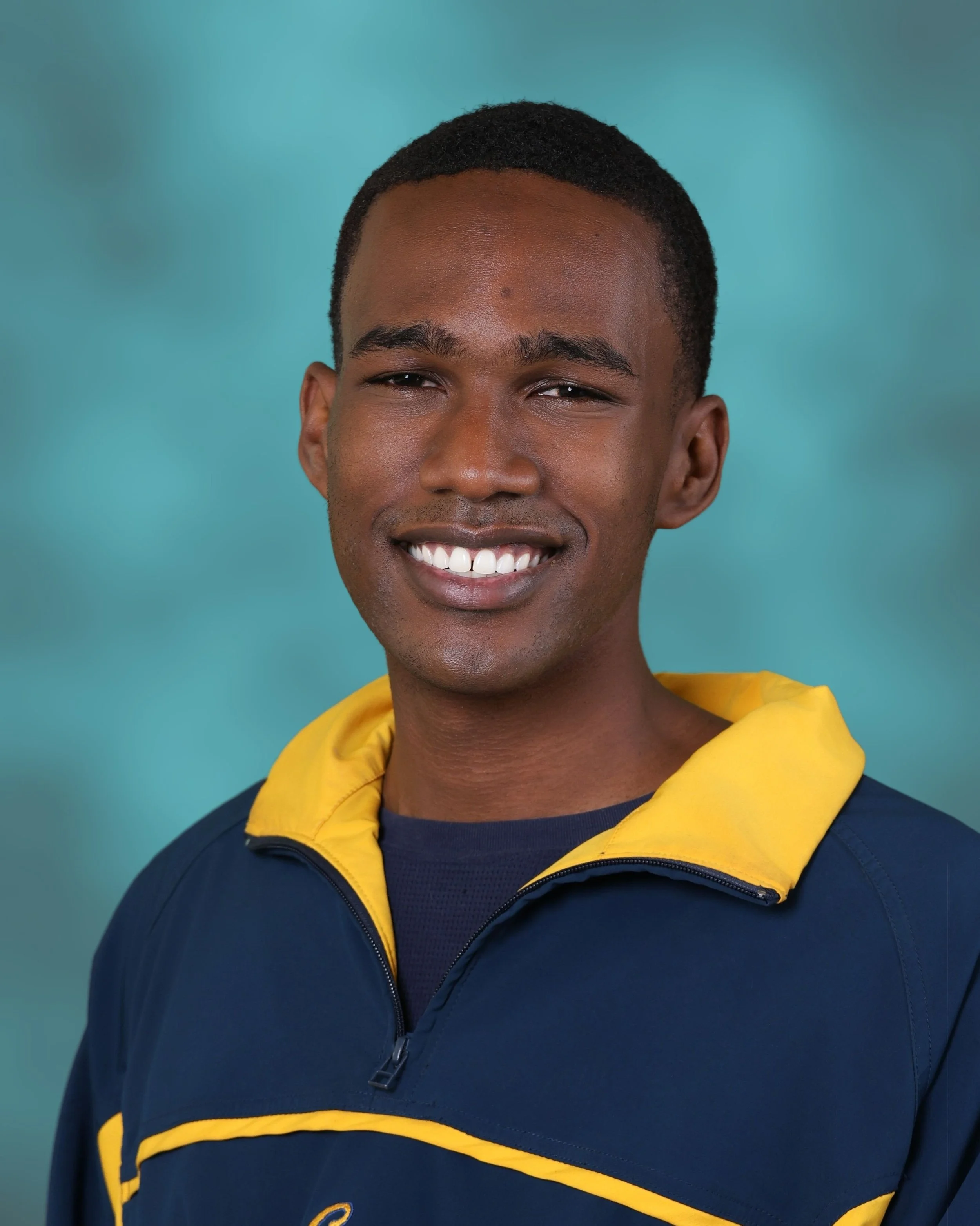 Close-up of a smiling young man with dark hair, wearing a blue jacket with yellow lining, against a blue background.