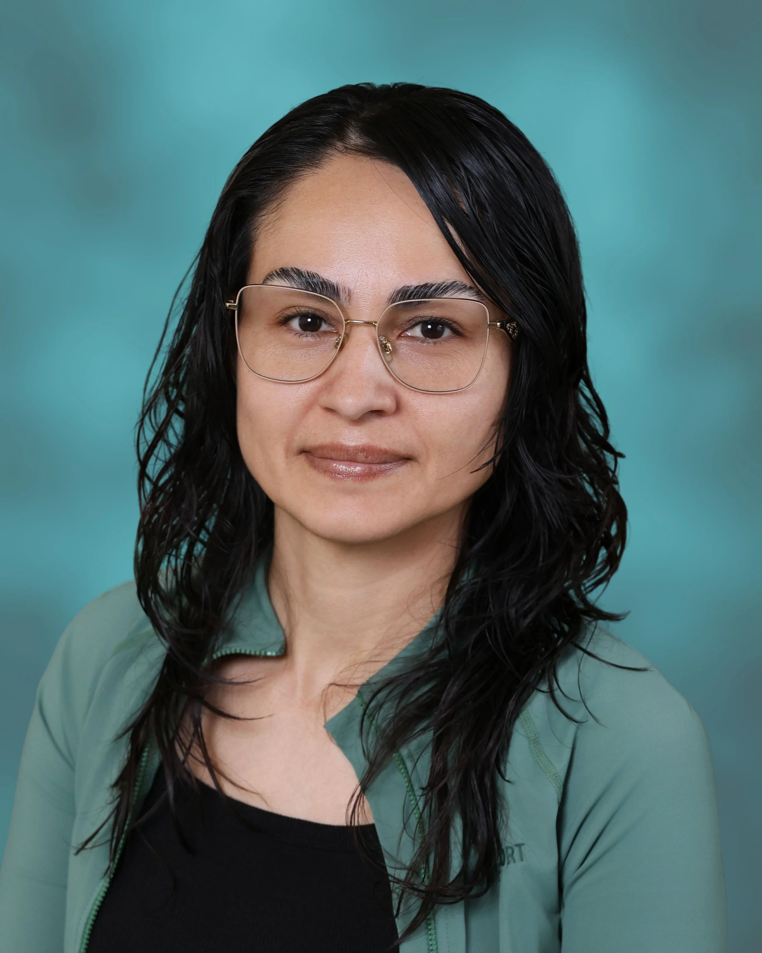 Close-up of a woman with black, wet, wavy hair, wearing glasses, a black top, and a light green jacket, smiling softly against a turquoise background.