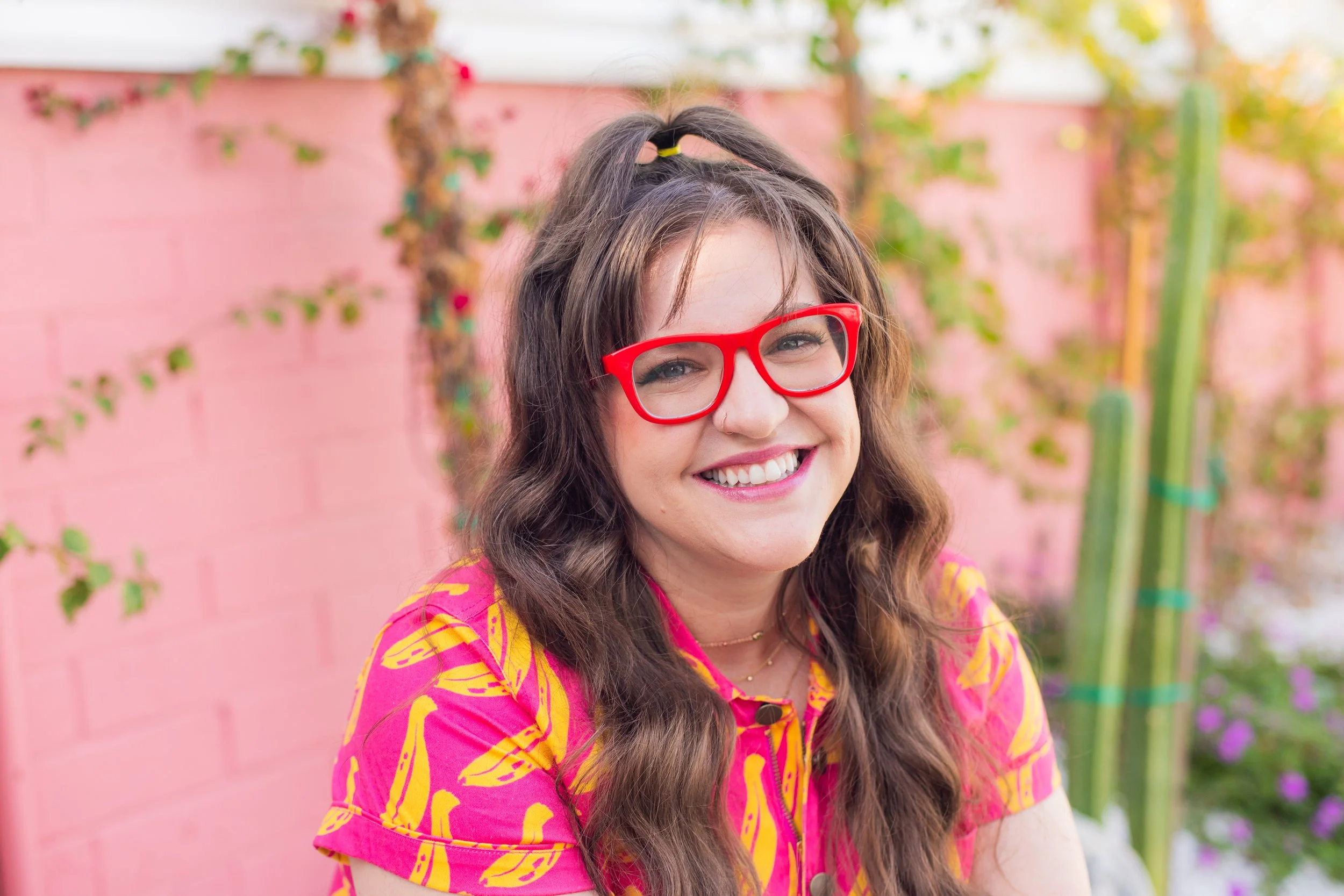 Smiling woman with red glasses wearing a colorful shirt in front of a pink wall with plants.