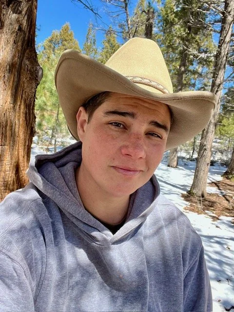 Flint in a tan cowboy hat and gray sweatshirt, slightly squinting at the camera. He has brown hair, dark eye brows, and brown eyes.