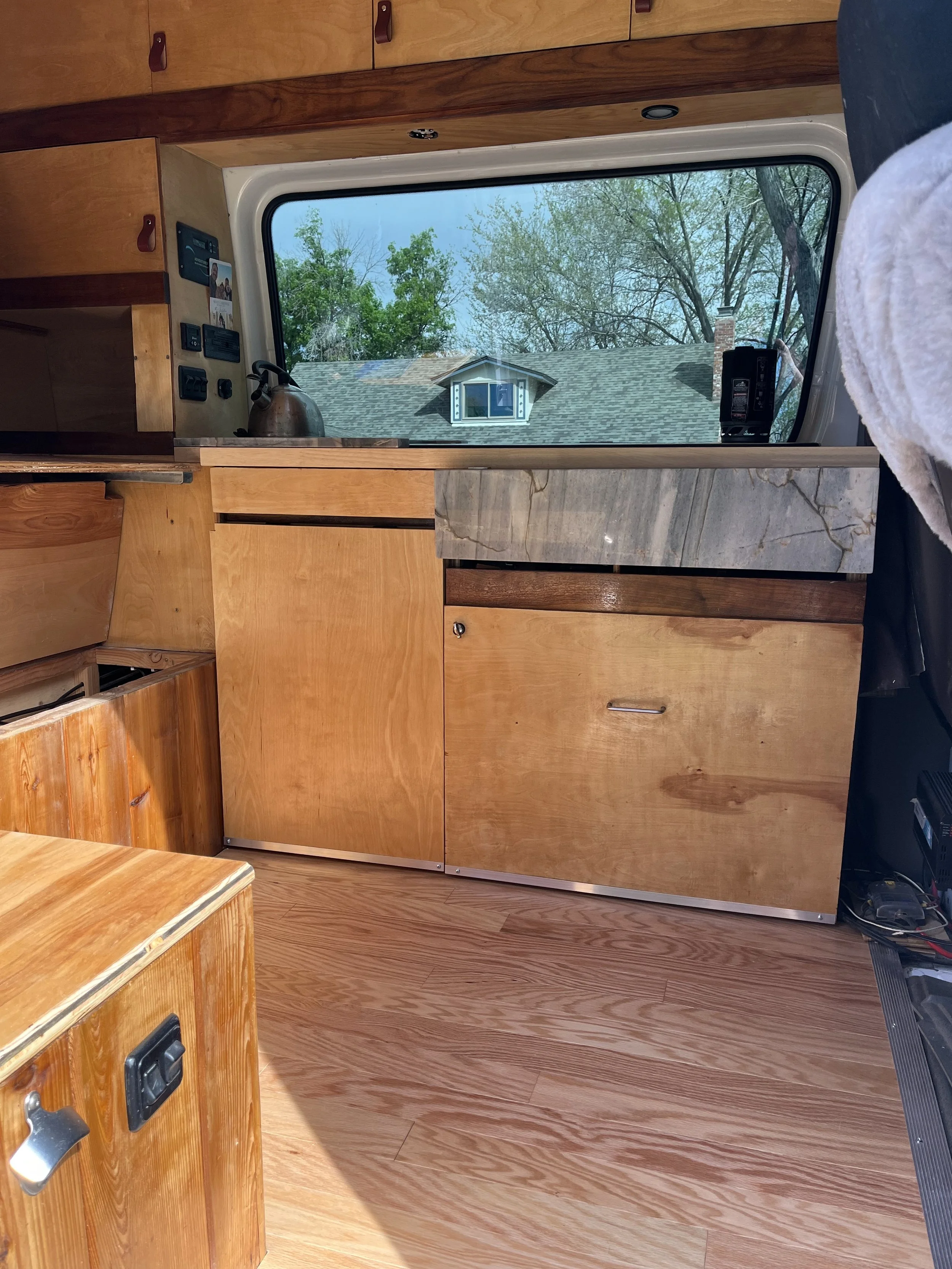 Interior view of a wooden kitchenette inside a camper van, with a window showing trees and a house roof outside.